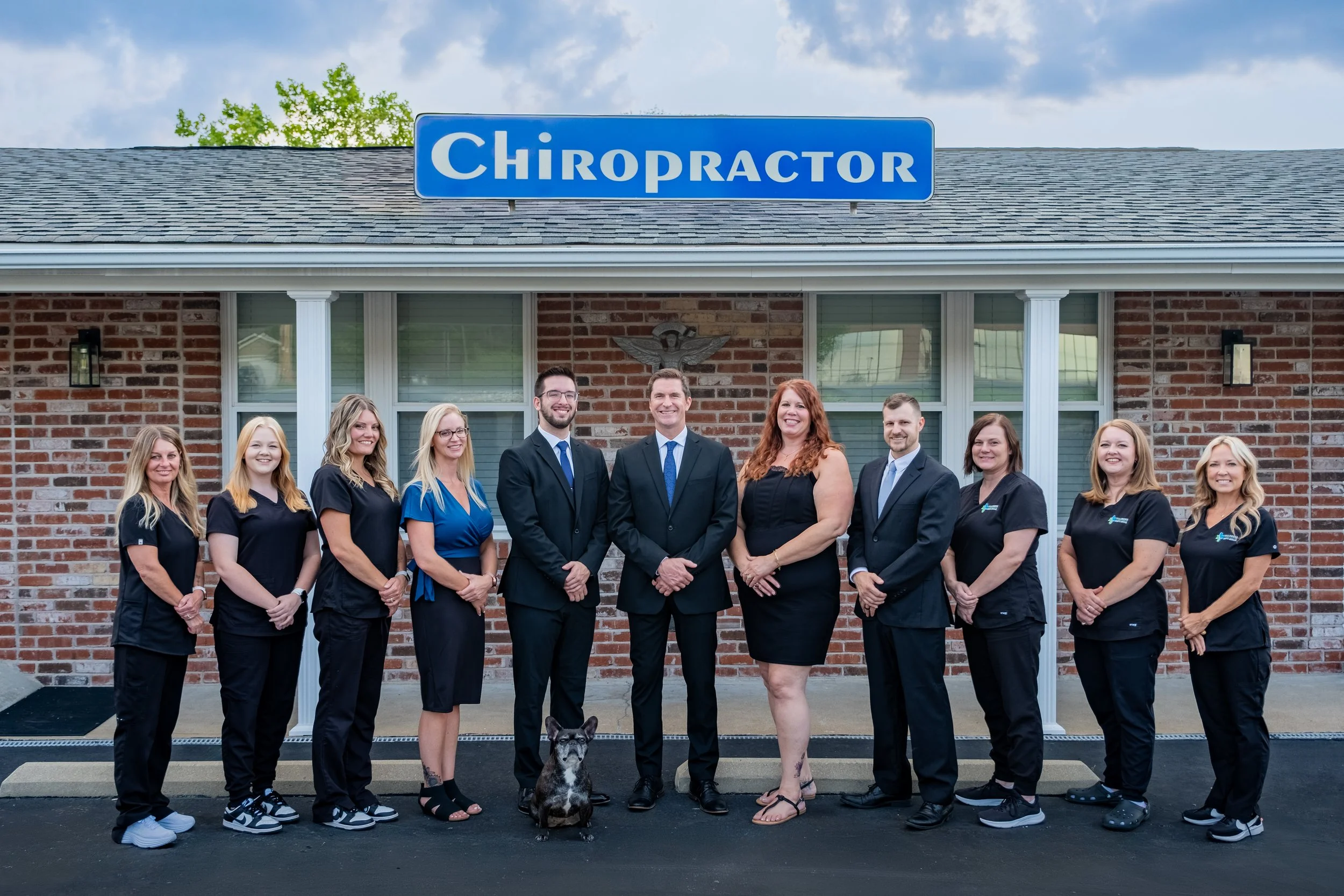 Group of ten people and a dog standing outside a brick building with a 'Chiropractor' sign on the roof