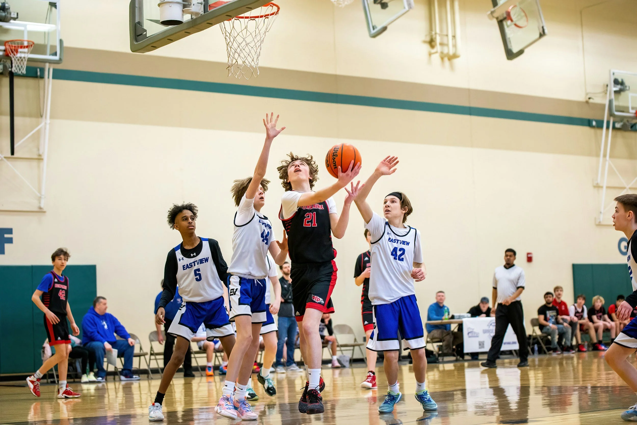 Young boy in black and red basketball uniform jumping to make a shot in an indoor gymnasium, surrounded by other children and a referee.