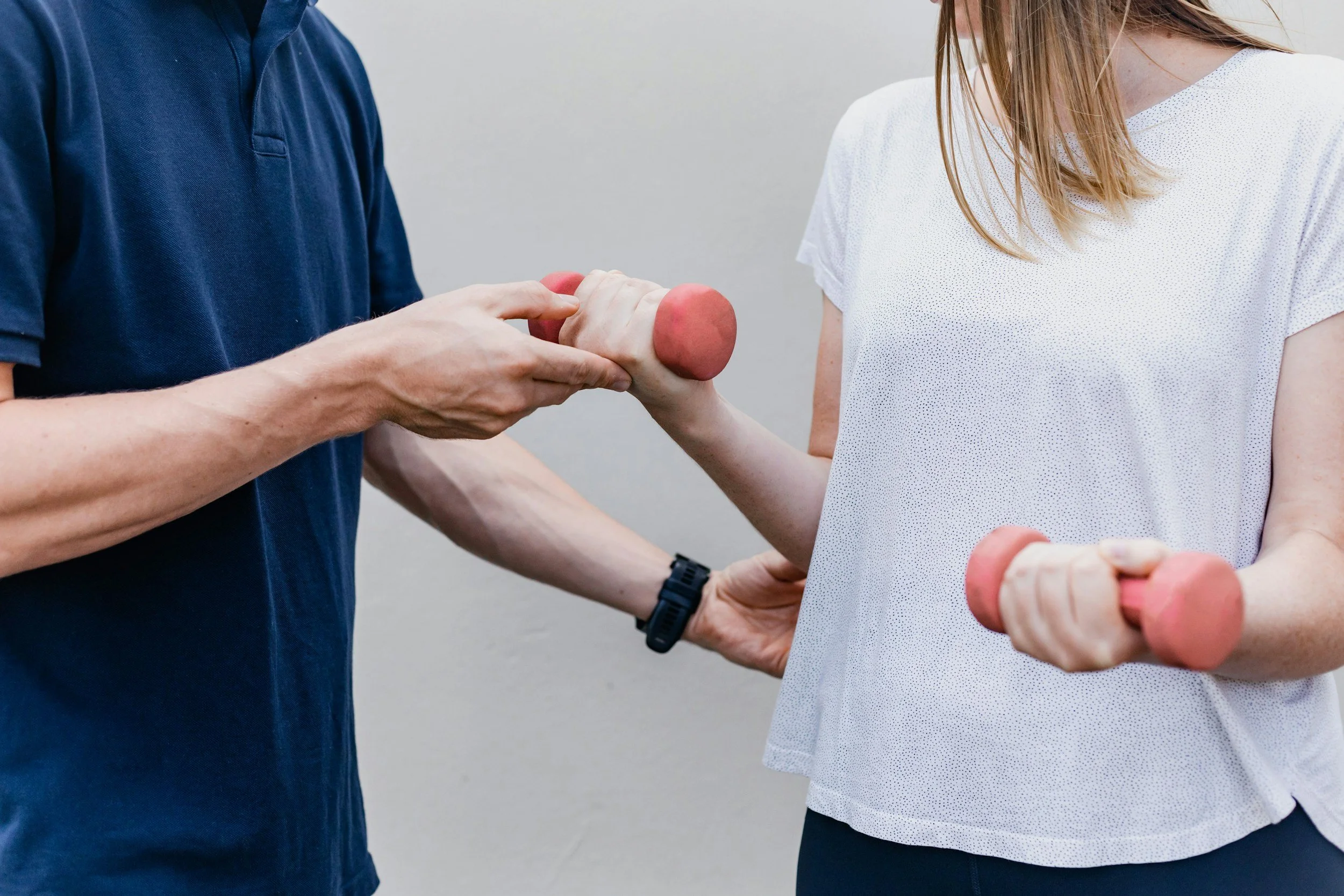A woman holding a pink dumbbell in her hand while a man, wearing a navy blue shirt, assists her in gripping it.