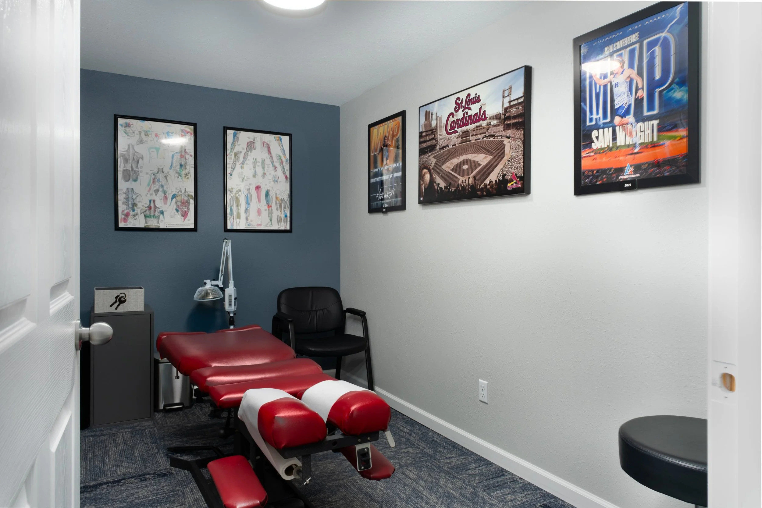 A chiropractic clinic room with a chiropractic adjustment table, a black chair, framed sports posters on the walls, and a grey cabinet with a tissue box on top.