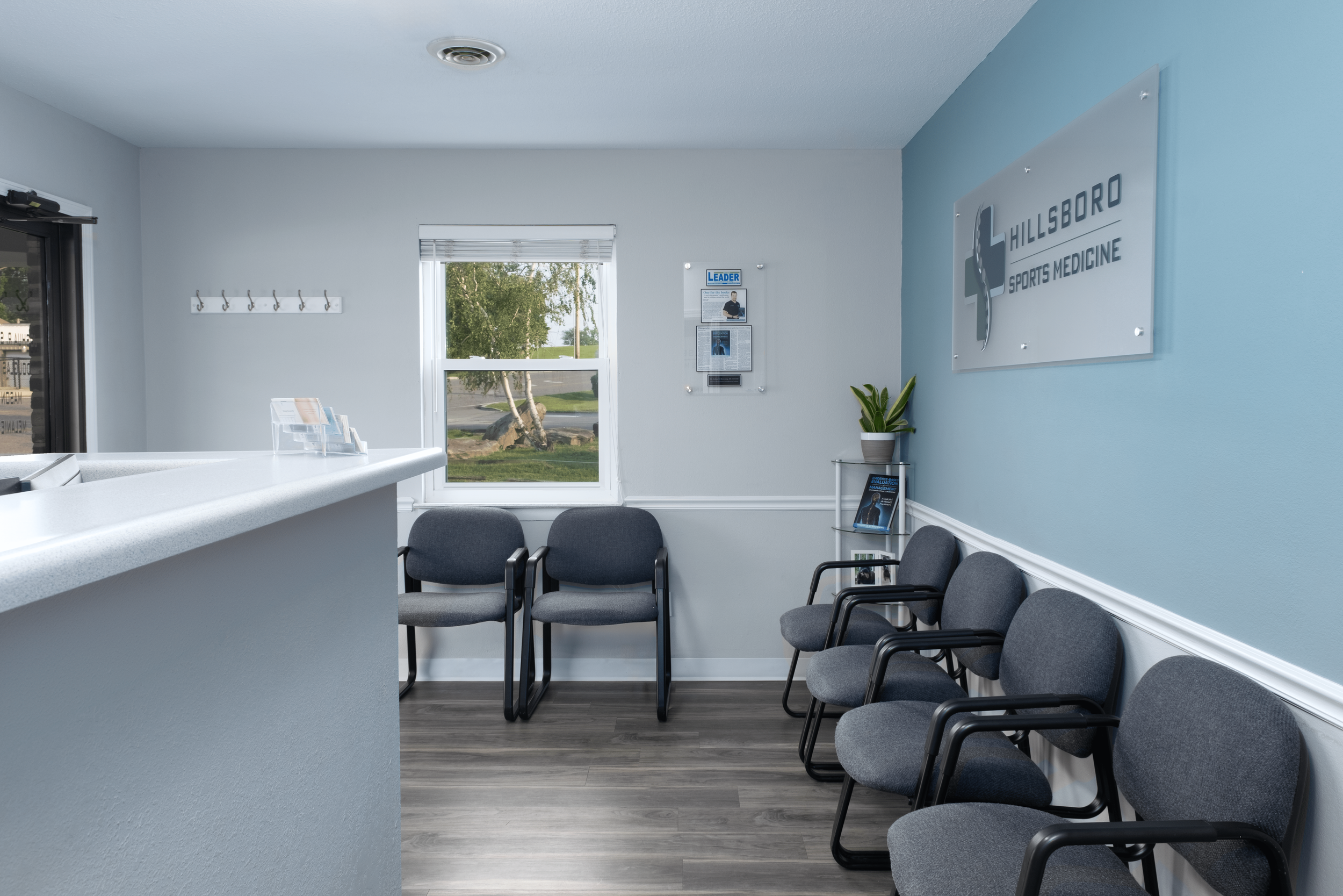 Empty waiting room at Hillsboro Sports Medicine with chairs arranged along the walls, a window showing greenery outside, and a sign on the wall.