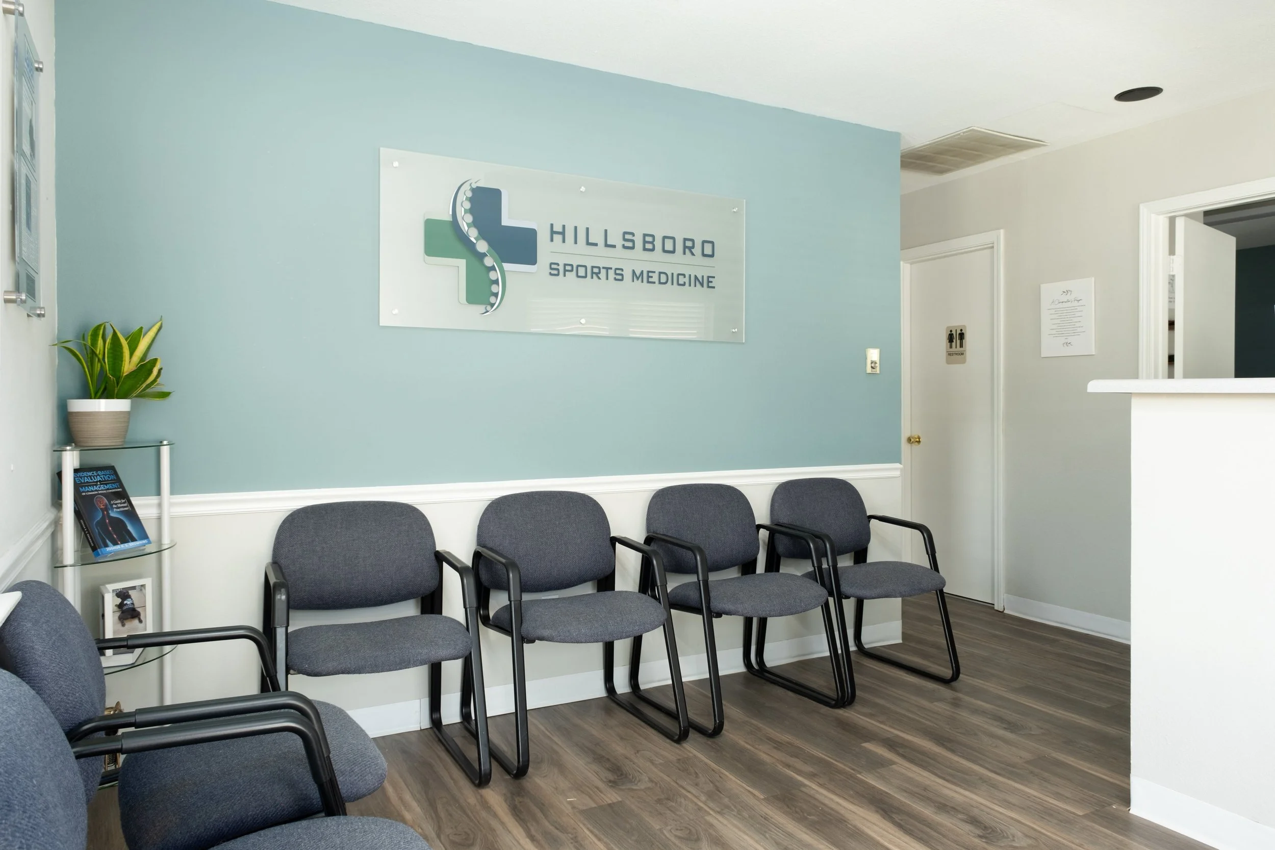 Waiting area in Hillsboro Sports Medicine clinic with five gray chairs, a potted plant, and a sign on the wall.