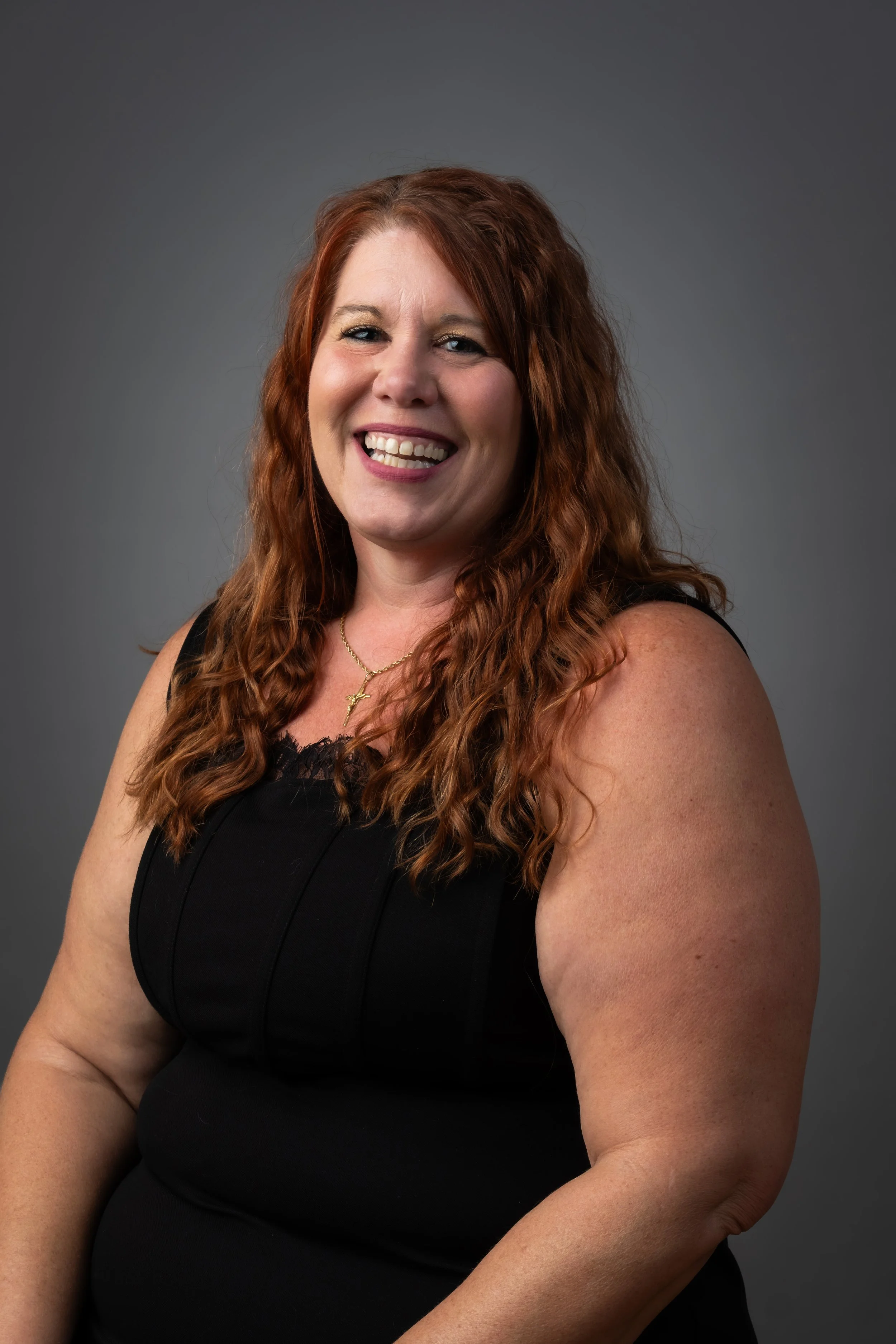 A woman with long curly red hair smiling, wearing a black sleeveless top, against a gray studio background.