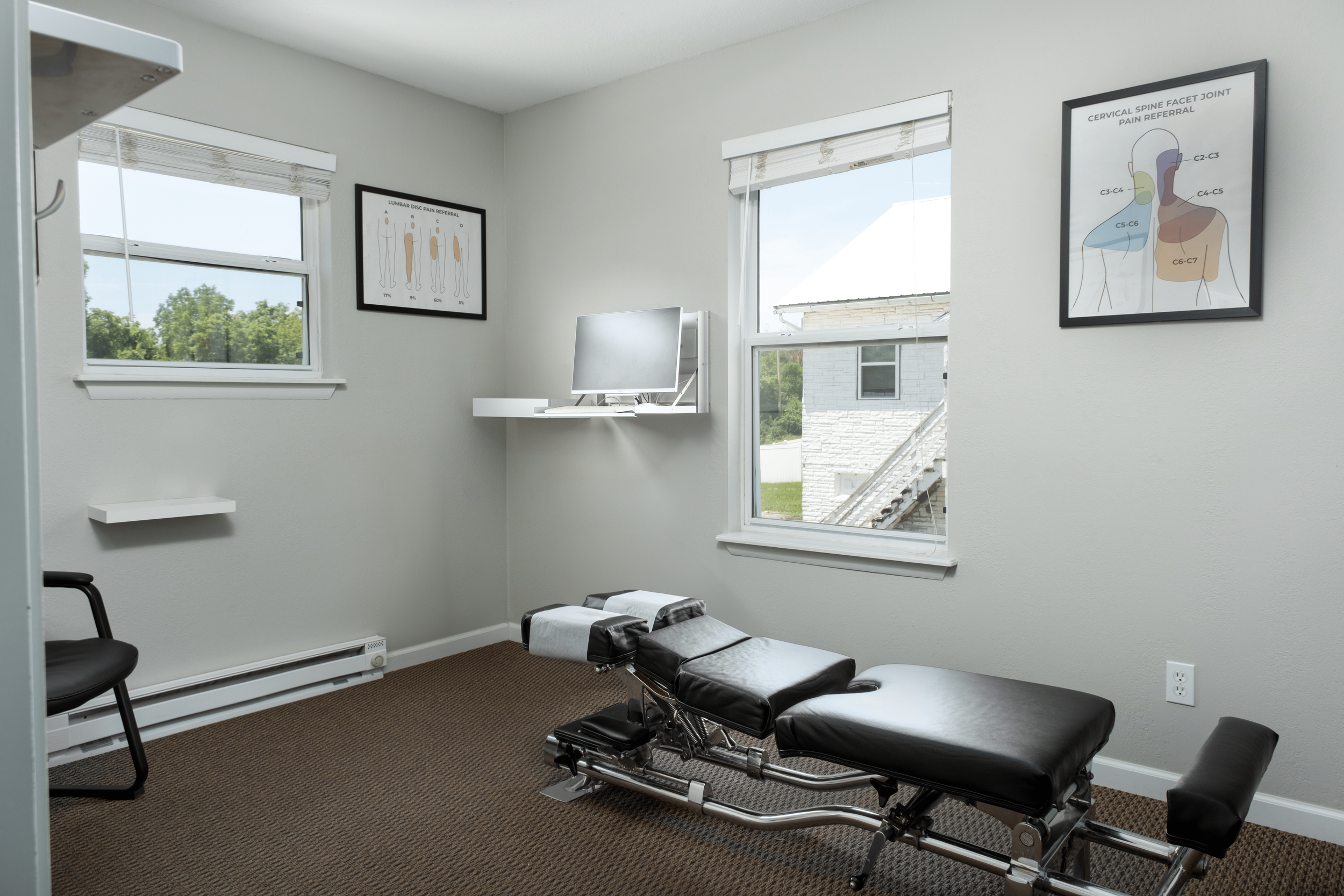 Medical examination room with a chiropractic treatment table, two windows, and posters of spinal anatomy and pain referral patterns on the wall.