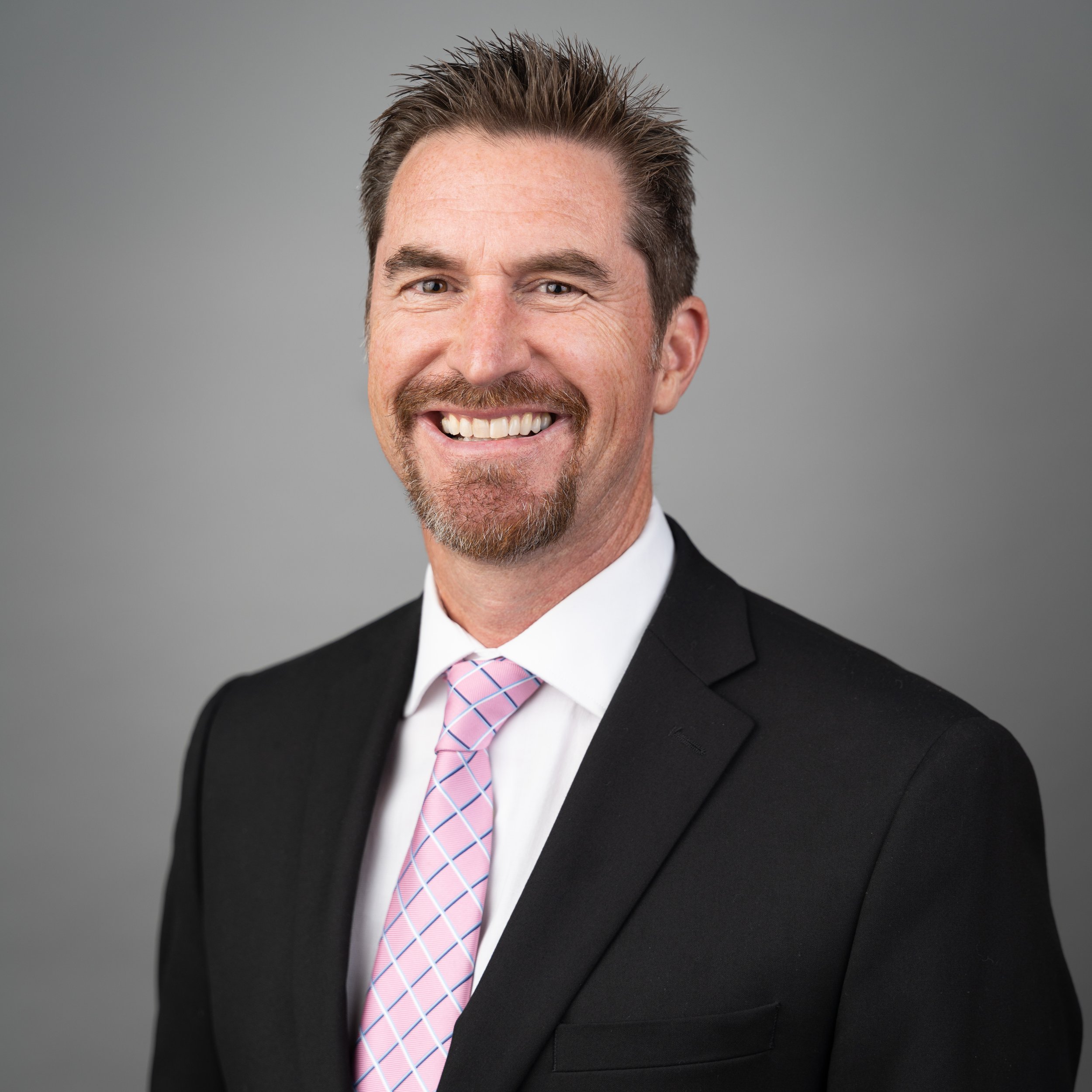 Headshot of a smiling man in a black suit, white shirt, and pink checkered tie, against a gray background.