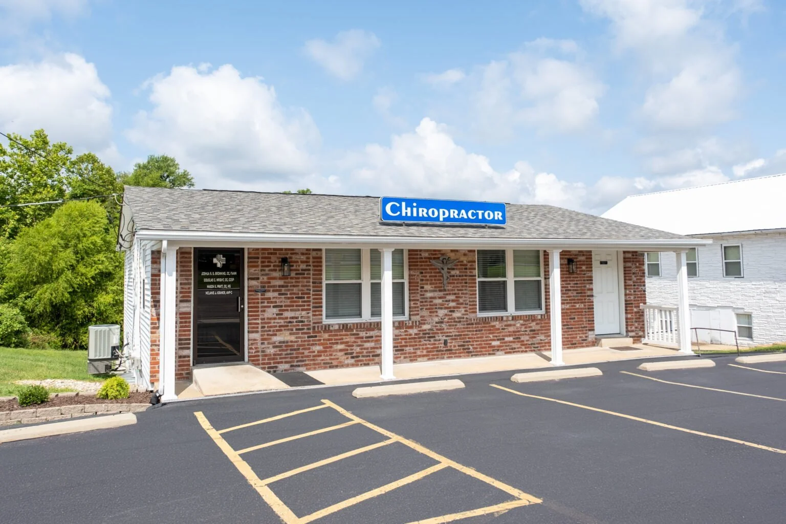 Brick building with a blue sign that reads 'Chiropractor', parking lot, and blue sky with clouds.