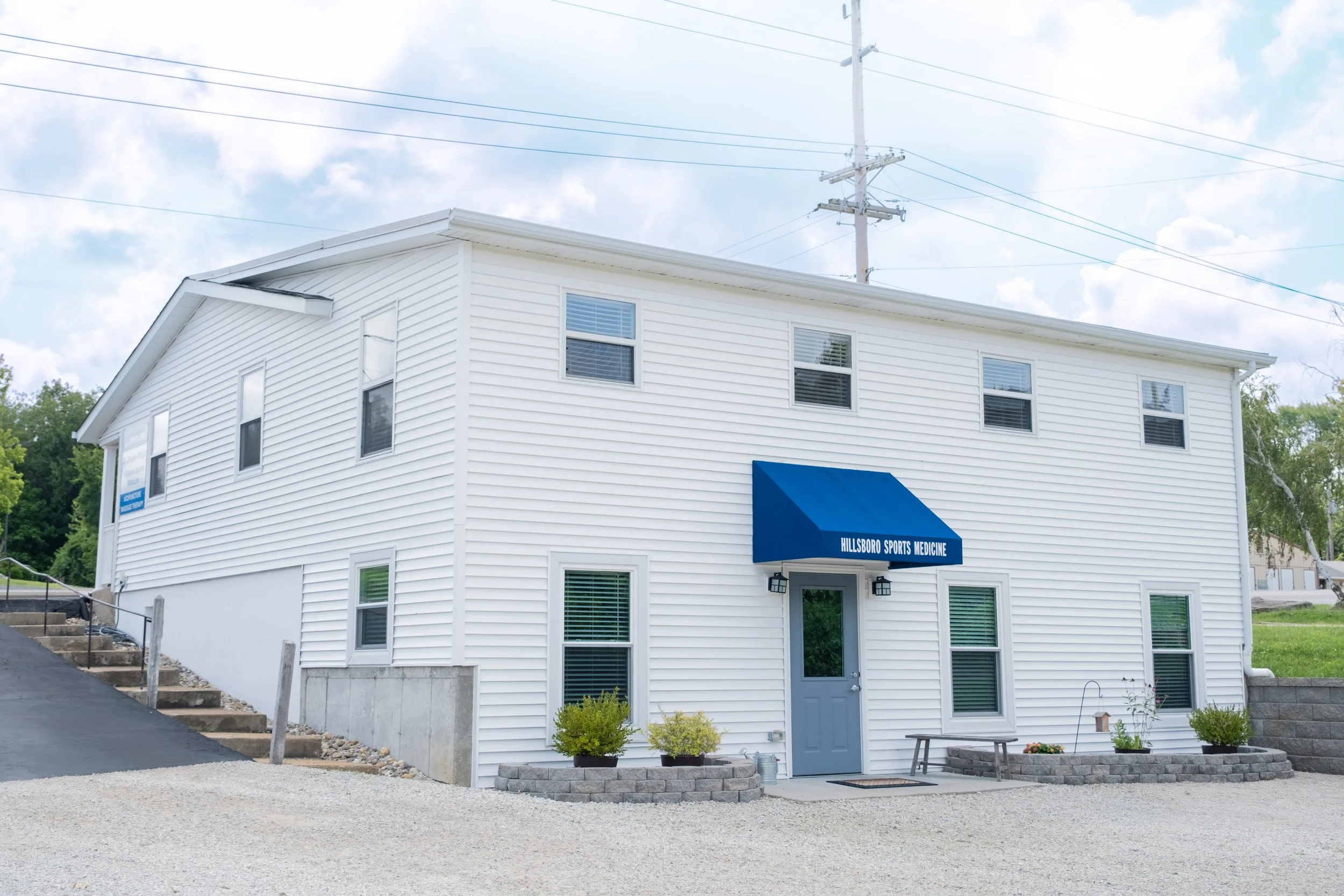 White two-story building with blue awning reading 'Hillsboro Sports Medicine', small front yard with potted plants, situated next to a paved driveway and gravel area, with overhead power lines and a partly cloudy sky.