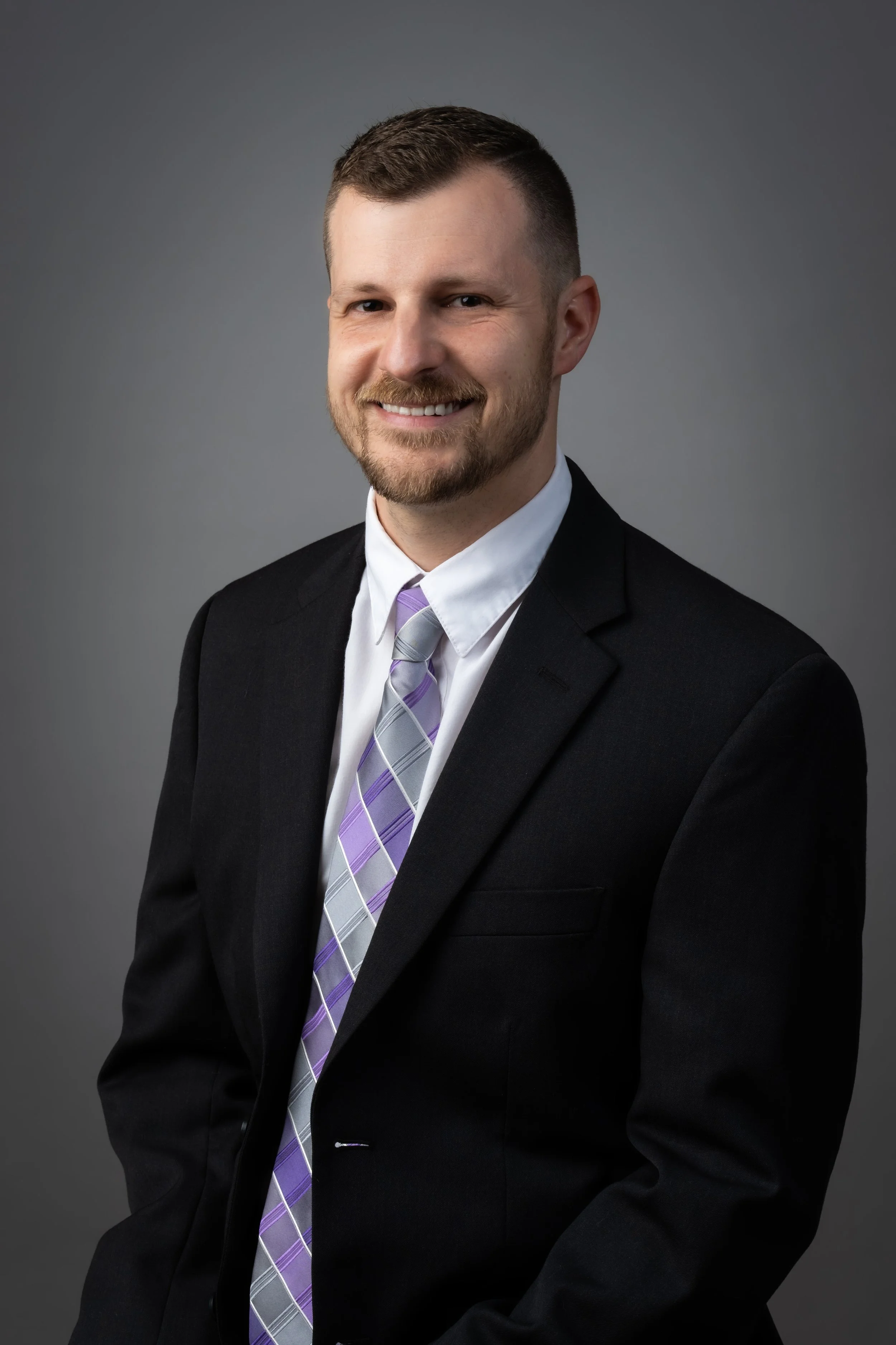 A professional man smiling, wearing a black suit, white shirt, and a purple double-striped tie, against a gray background.