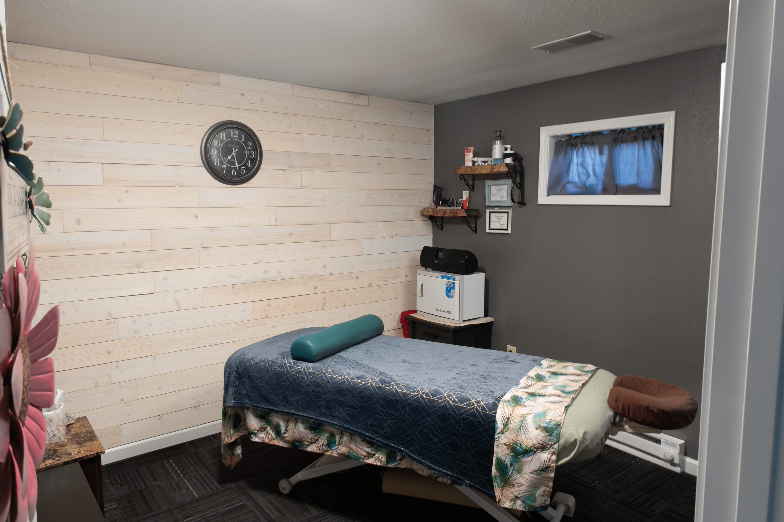 A massage therapy room with a massage bed covered with a patterned blanket and a bolster pillow, a clock on a wood-paneled wall, and shelves with various items on a gray wall.