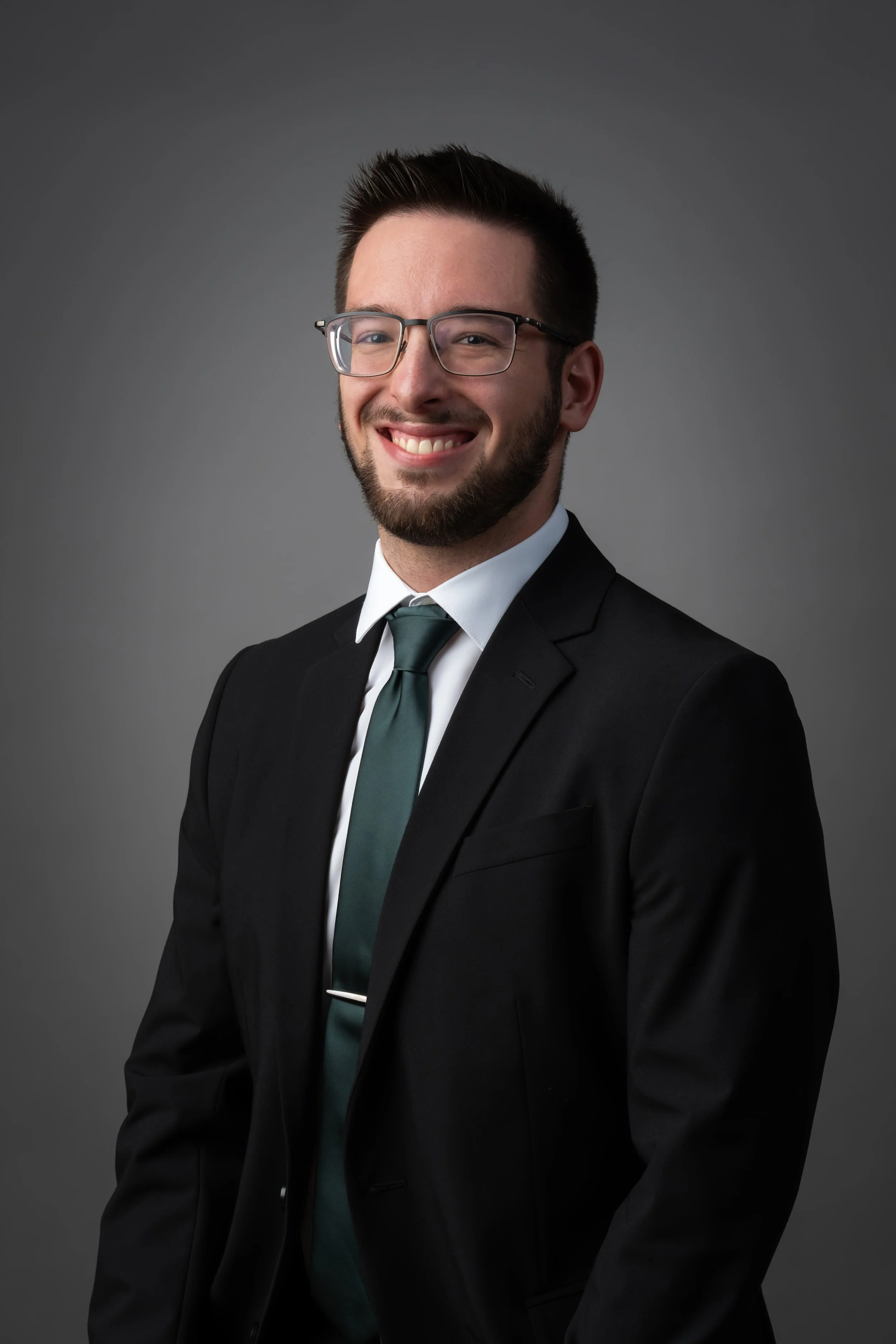 A young man in a black suit, white shirt, and teal tie, smiling with glasses, posed against a gray background.