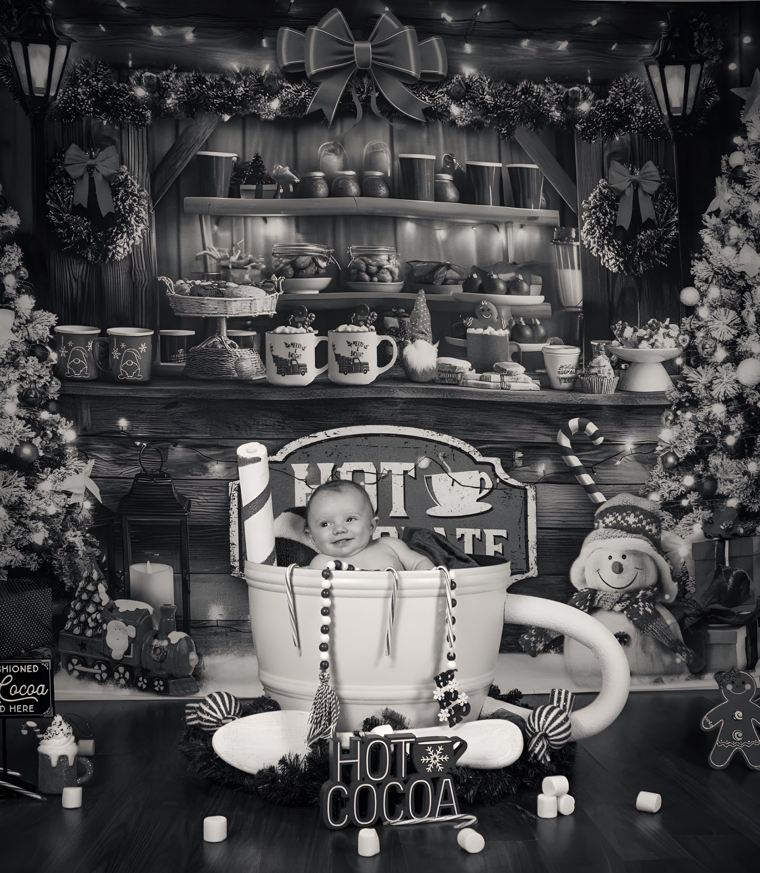 A baby sitting inside a large coffee cup prop for a festive holiday photo, surrounded by Christmas decorations including a decorated Christmas tree, a snowman plush, and holiday candies. The background features a holiday-themed retail display with baked goods, mugs, and Christmas wreaths.