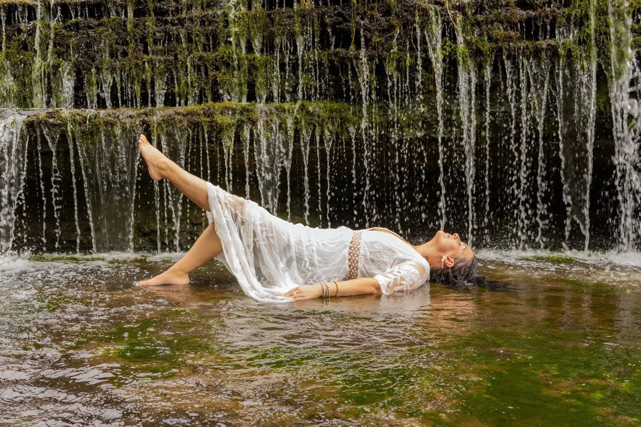 A woman in a white dress lying on her side in a shallow stream, with a waterfall in the background.