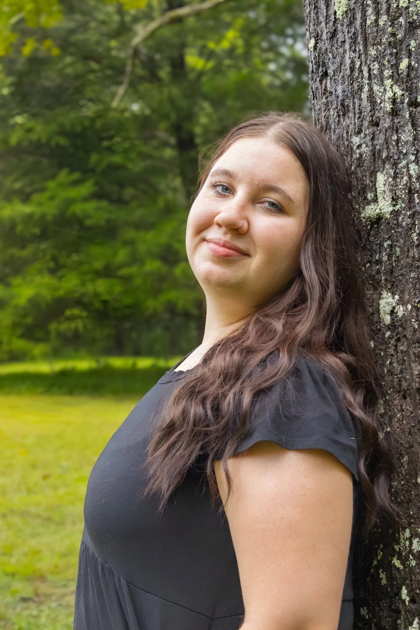 Young woman with long wavy hair leaning against a tree in a green park, smiling softly.