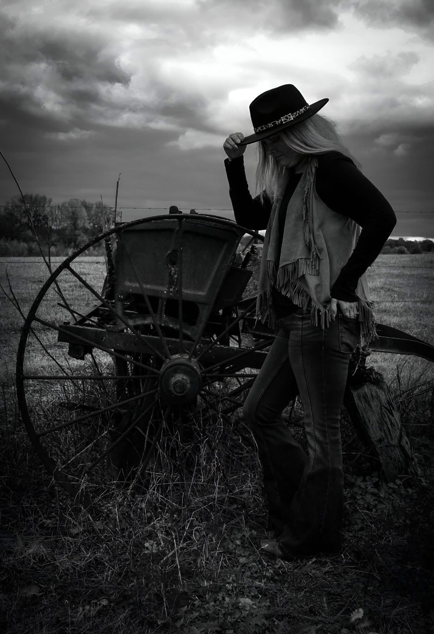 A woman wearing a wide-brimmed hat and fringed vest stands outdoors near an old farming machine under a cloudy sky.