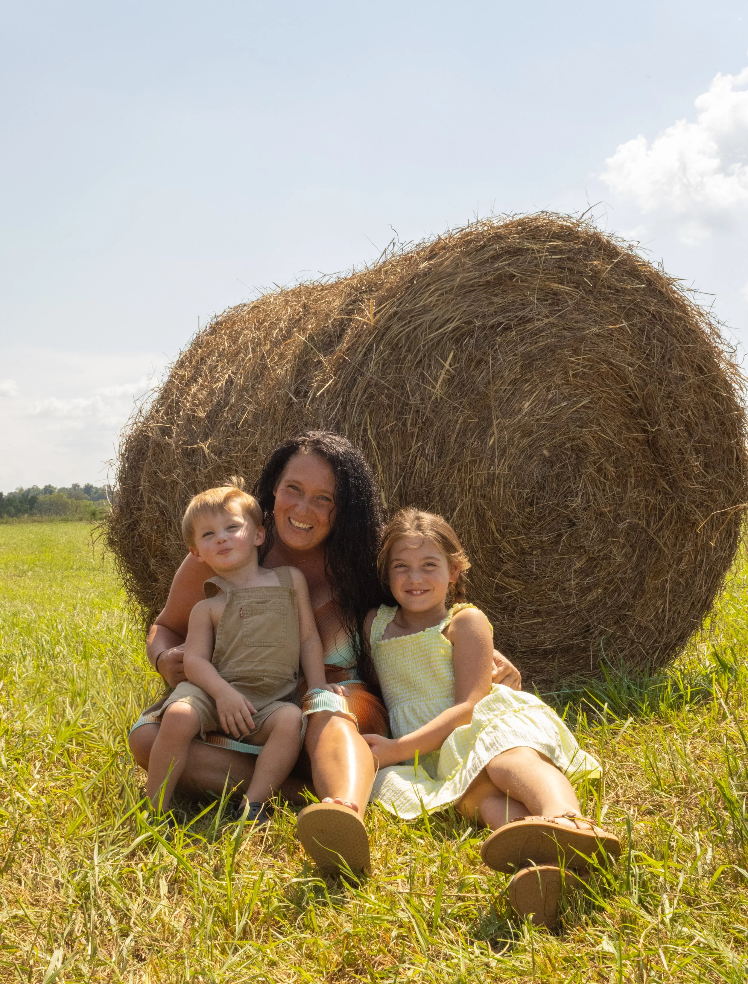 A woman and two children sitting on the grass in front of a large hay bale in a field, smiling at the camera.