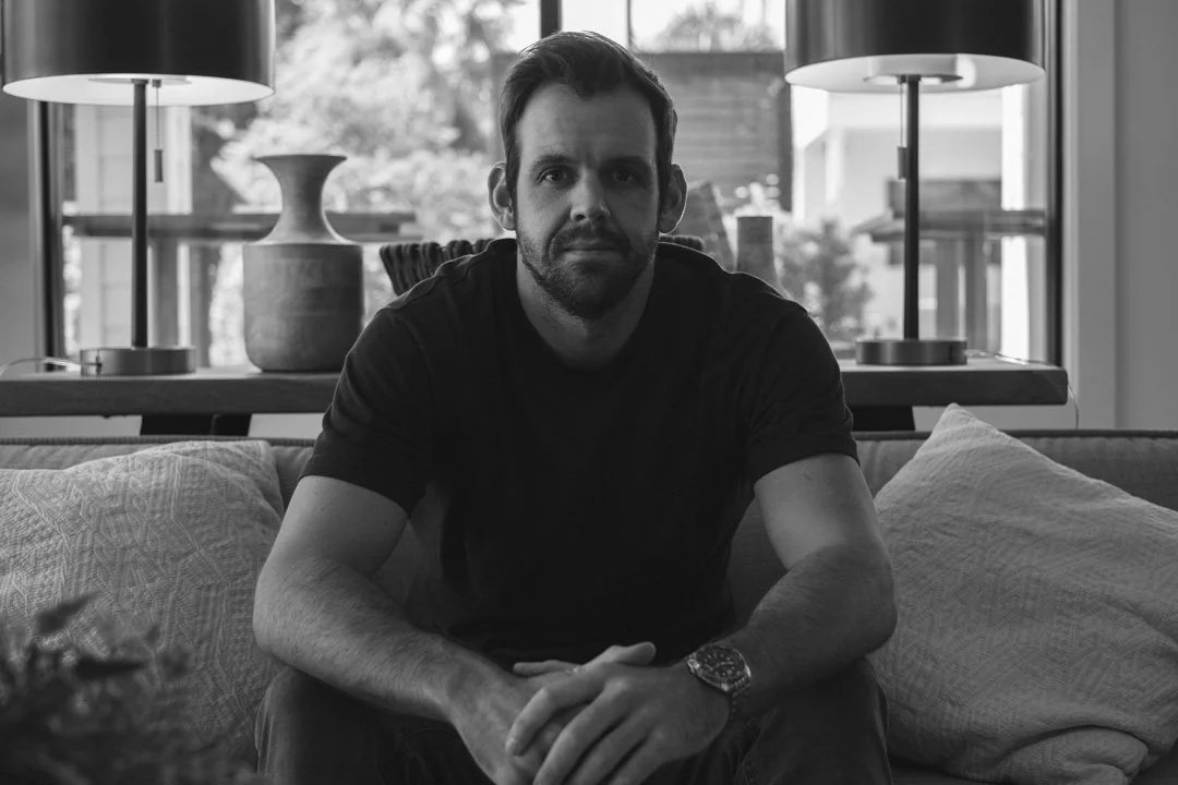A man with a beard and short hair sitting on a couch in a living room, with two lamps, a large vase, and windows in the background, in black and white.