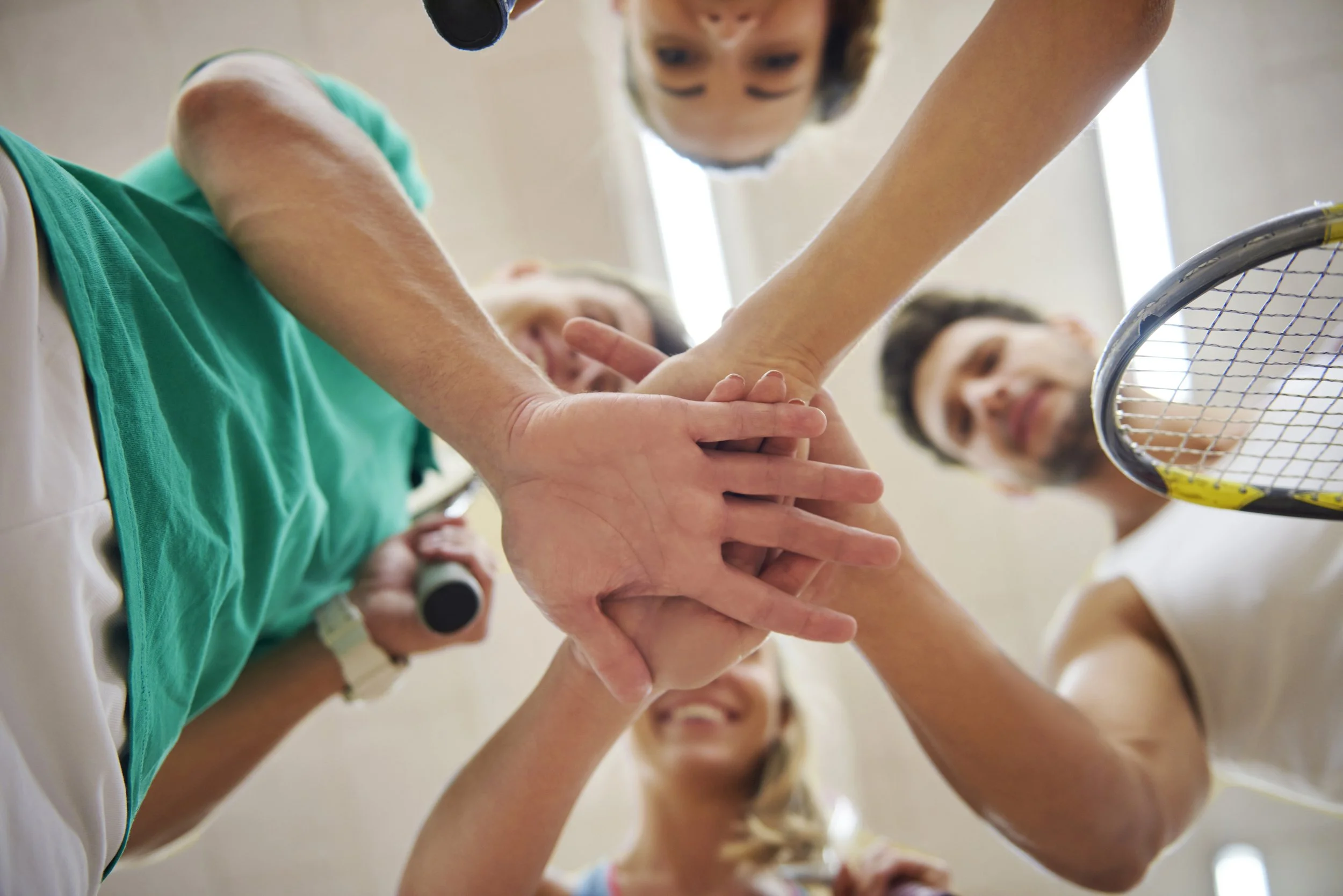 Four people, including children and a man with a racket, putting their hands together in a huddle with smiles, as if preparing for a game.