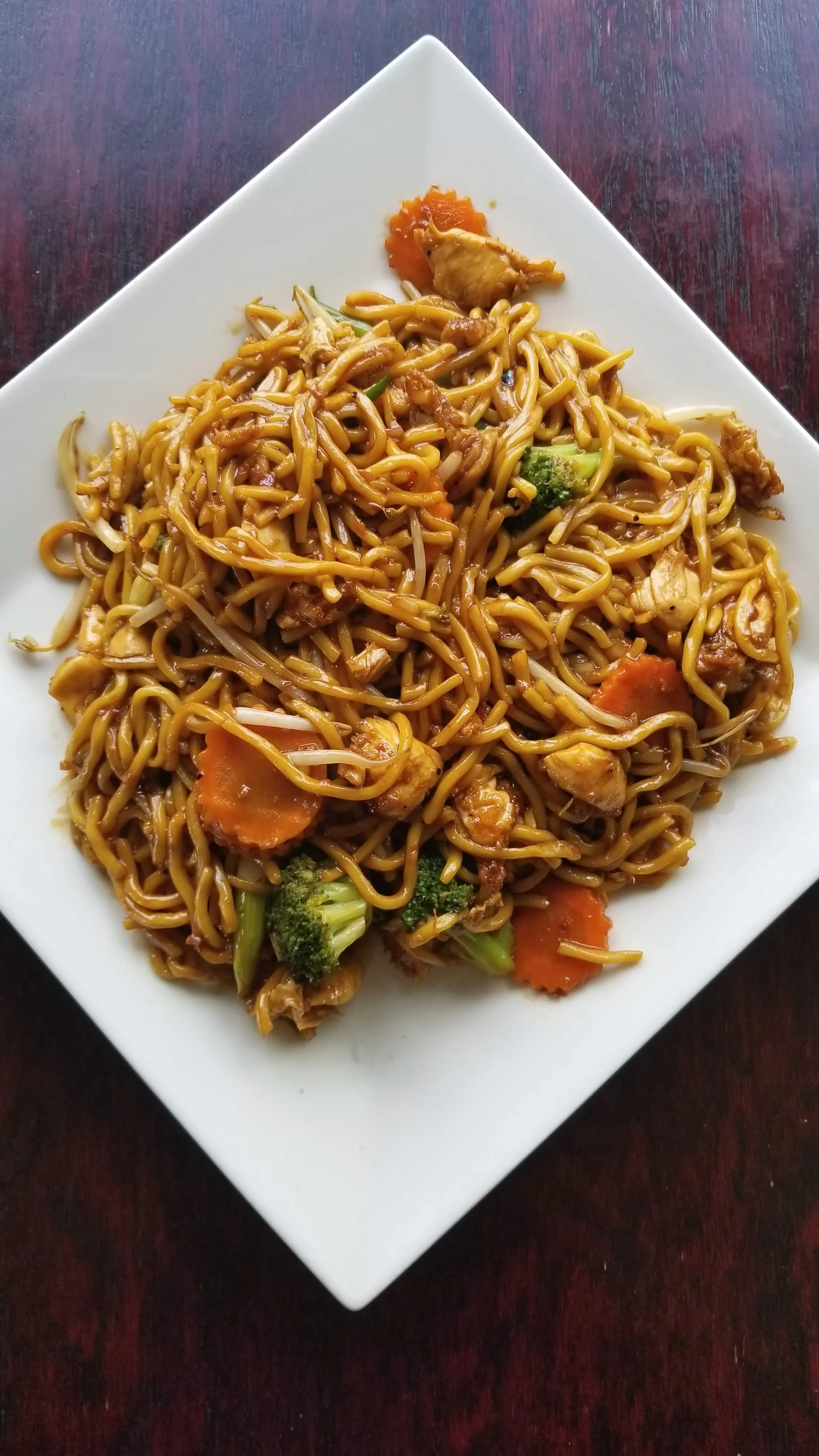Plate of stir-fried noodles with vegetables on a white square plate. Includes broccoli, carrots, and chicken pieces.
