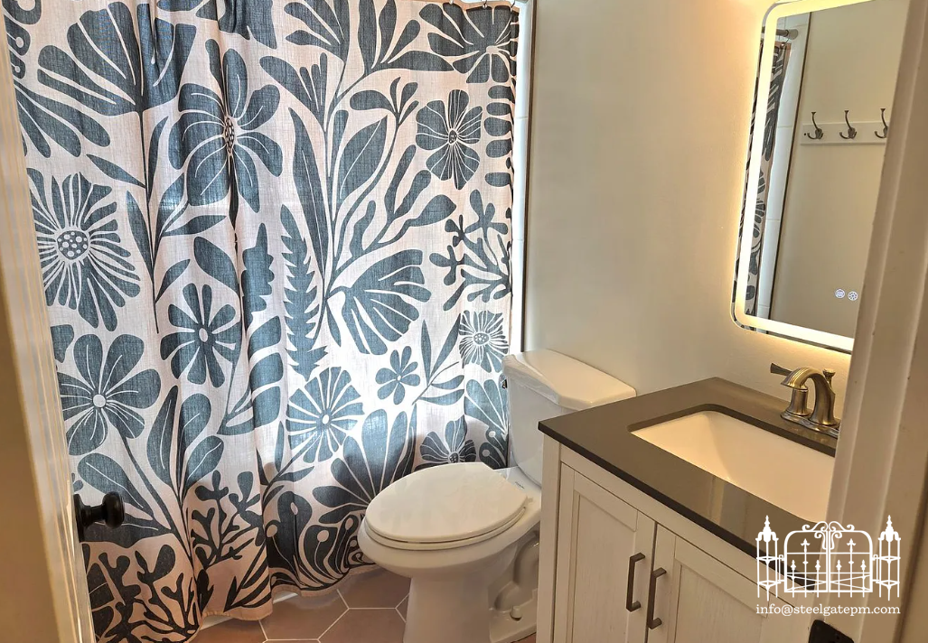 Bathroom with a floral shower curtain, toilet, white cabinet with a black countertop and under-mount sink, and a mirror with built-in lighting.