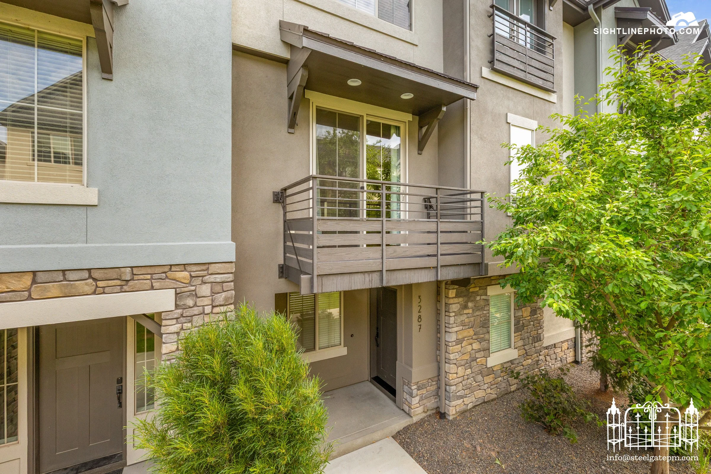 Exterior view of a modern apartment building with a small balcony, large windows, stone and stucco siding, a neighboring door, greenery, and a decorative white fence in the bottom right corner.