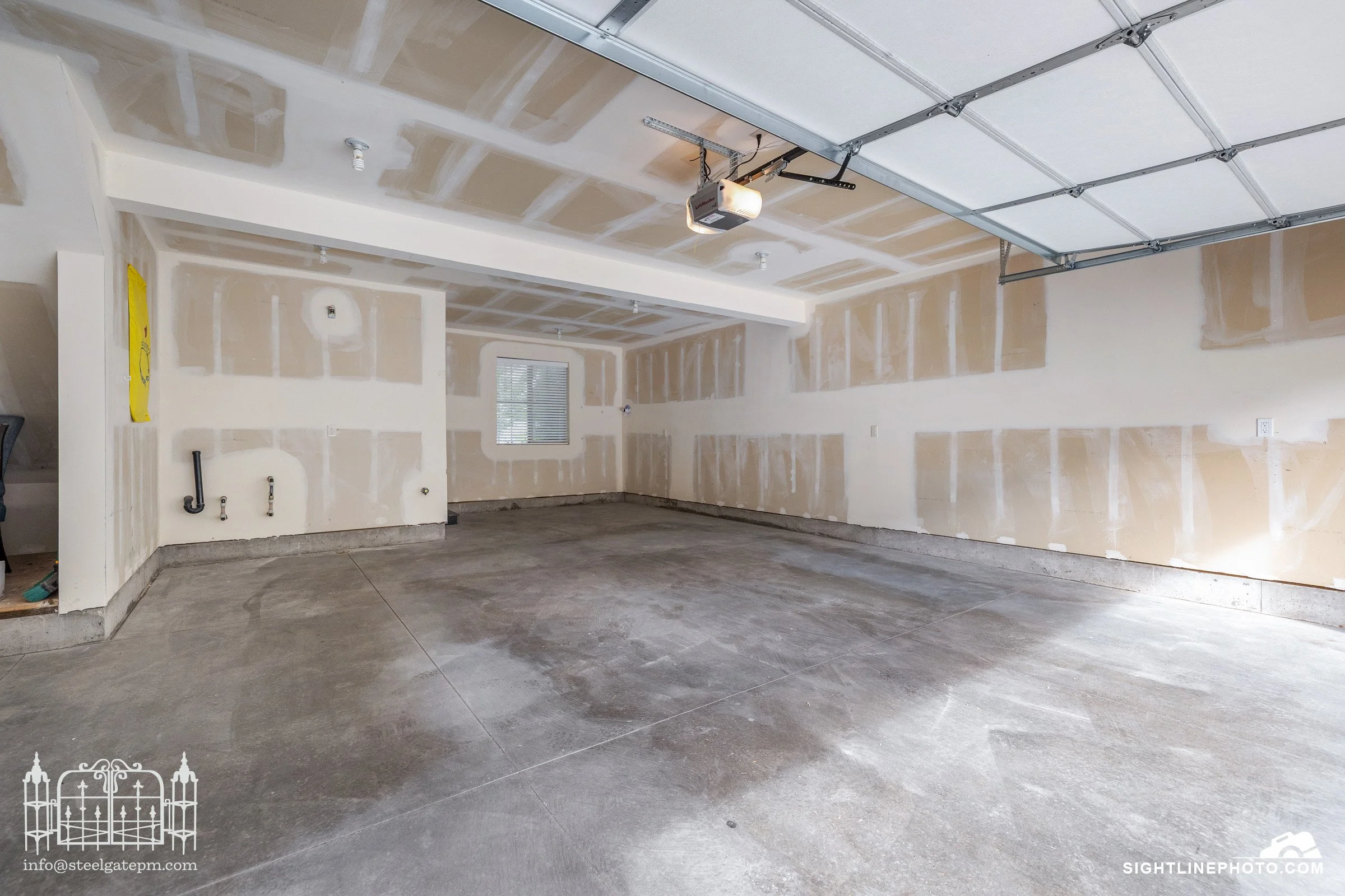 Empty garage with unfinished drywall and a concrete floor. Garage door is open, and a small window is on one wall. No vehicles or furniture inside.