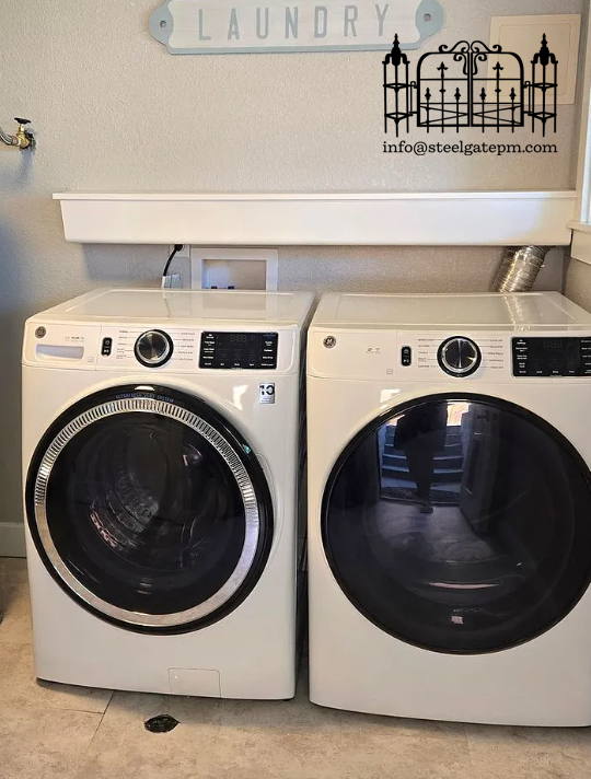 A laundry room with a front-loading washing machine on the left and a matching dryer on the right. Above them, there is a decorative sign that says 'LAUNDRY' and a black metal wall ornament. An email address is also visible on the wall.