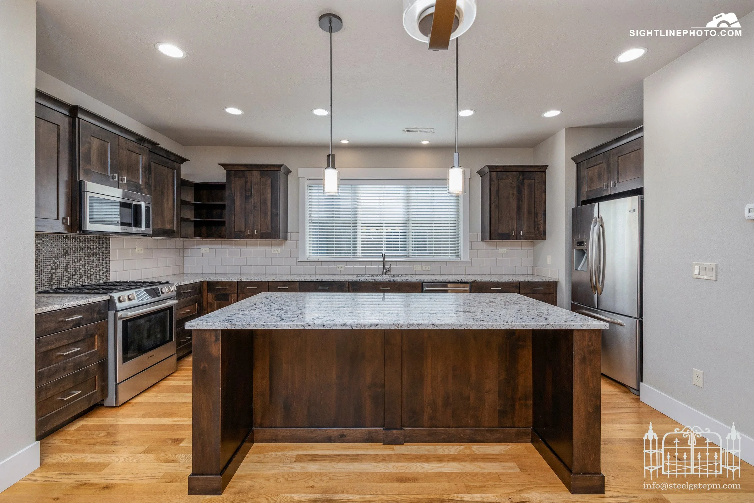 Modern kitchen with dark wooden cabinets, stainless steel appliances, granite countertops, a large center island, and a window with white blinds.