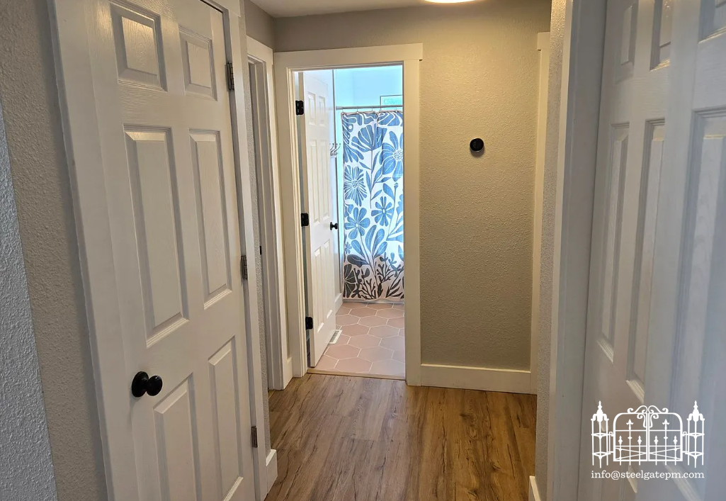 A hallway with a view into a bathroom with a patterned curtain, hardwood flooring, and white doors.