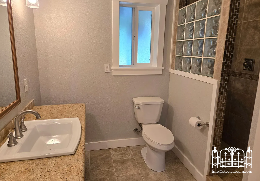 Bathroom with a granite countertop sink, a mirror, a toilet, a small window, and a glass block wall section separating the shower area.