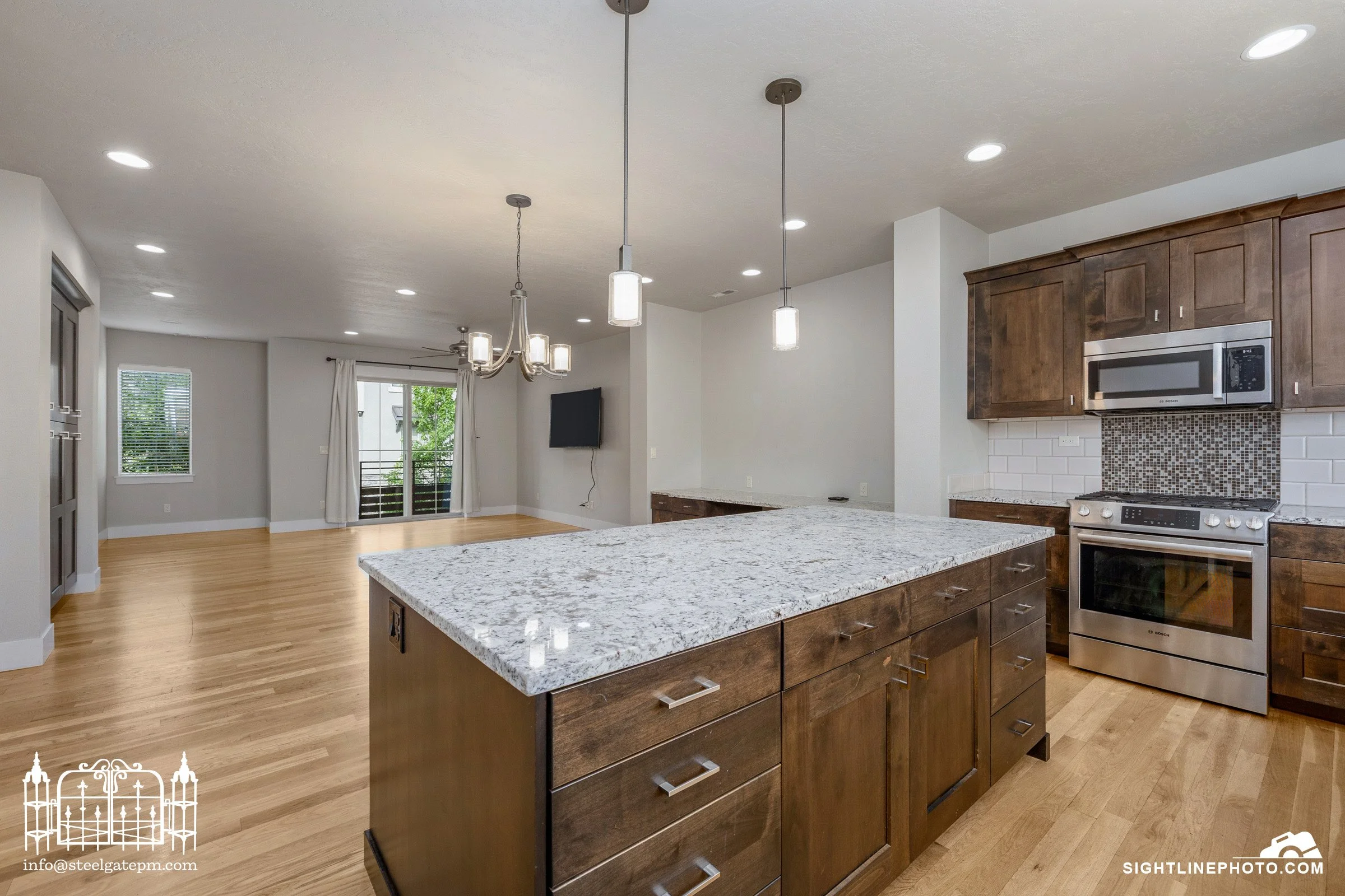 Open-concept kitchen with granite island, wooden cabinets, stainless steel microwave and stove, hardwood floors, and a chandelier over the dining area with sliding glass doors leading to a balcony.