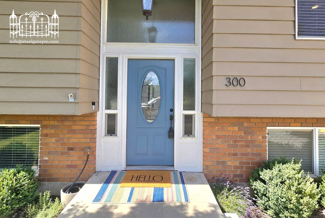 Front door of a house with a blue door, welcome mat reading 'HELLO', and house number 300. The house features beige siding and brick accents, with bushes and plants on either side.