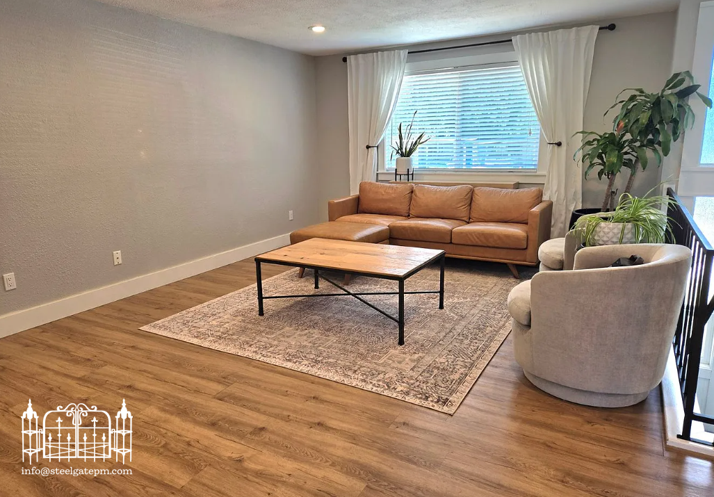 Living room with a tan sofa, white armchair, potted plants, large window with white curtains, wooden floor, and a rustic coffee table, minimal decor.