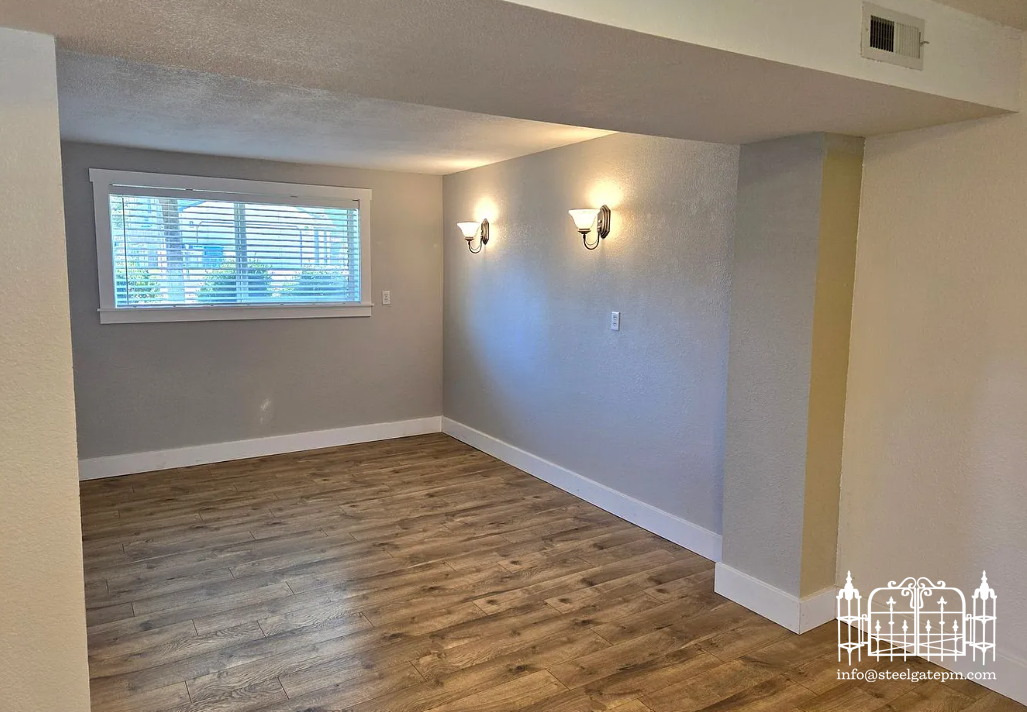 Un empty room with a window, wooden flooring, gray walls, and two wall-mounted light fixtures.