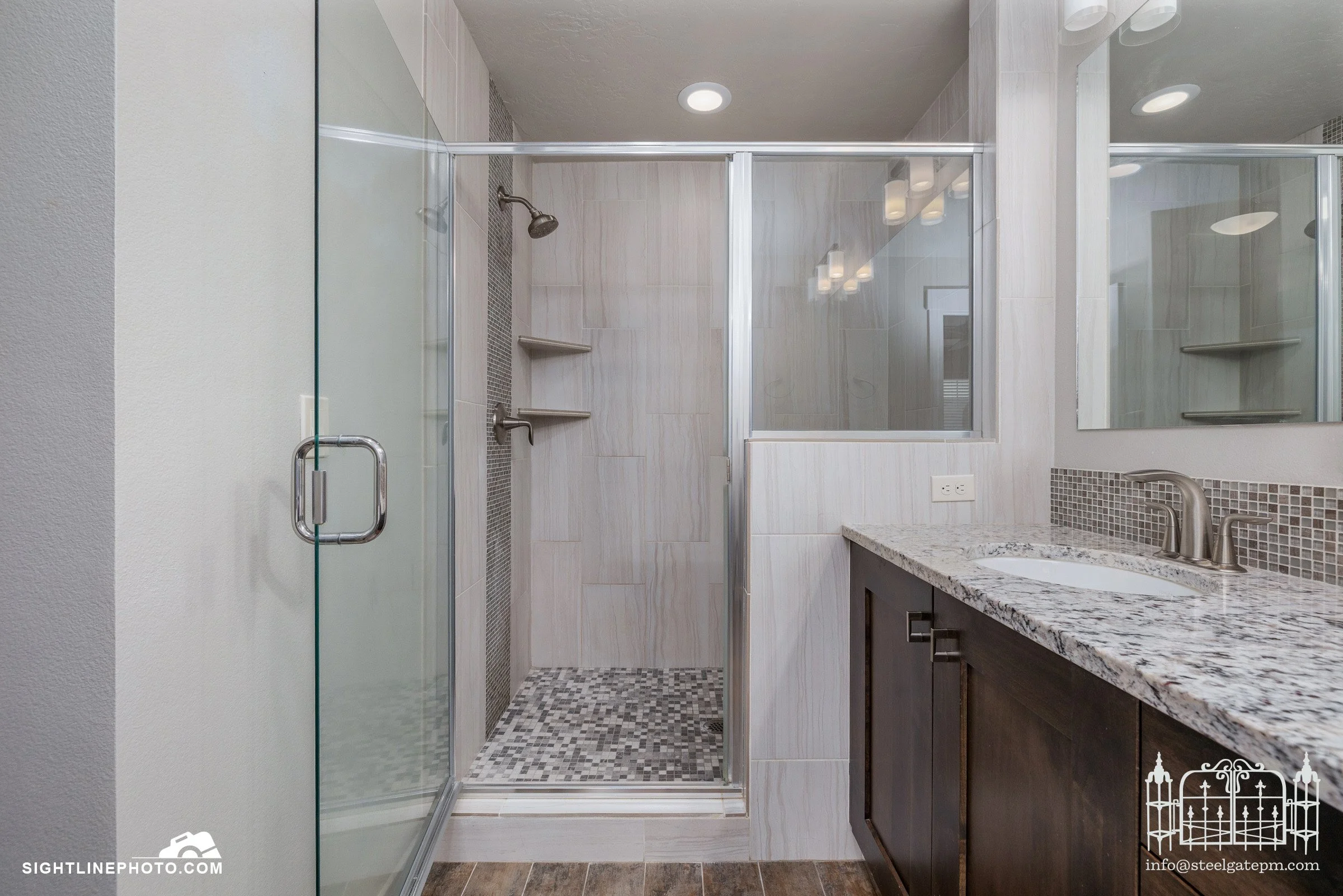 A modern bathroom with a glass-enclosed shower, a granite countertop sink, dark wood cabinets, and a large mirror.