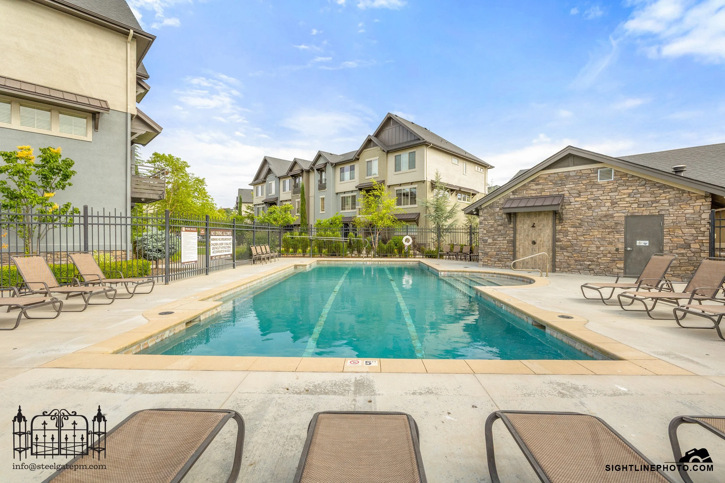 An outdoor swimming pool area with chairs around it, enclosed by a black metal fence. In the background, there are multi-story residential buildings with various architectural styles and a blue sky with scattered clouds.
