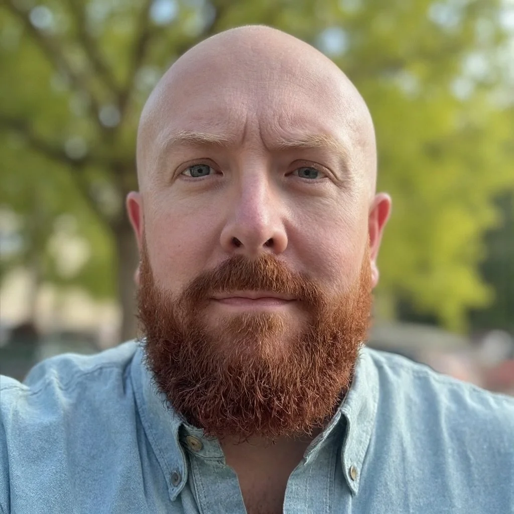 Close-up of a bald man with a full red beard, wearing a light blue button-up shirt, outdoors with green blurred trees in the background.