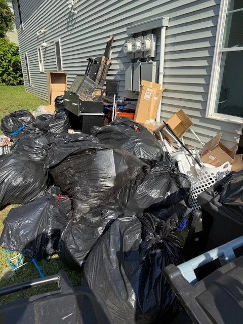 Overflow of trash bags and discarded items outside a house, near utility meters and the side of a building.