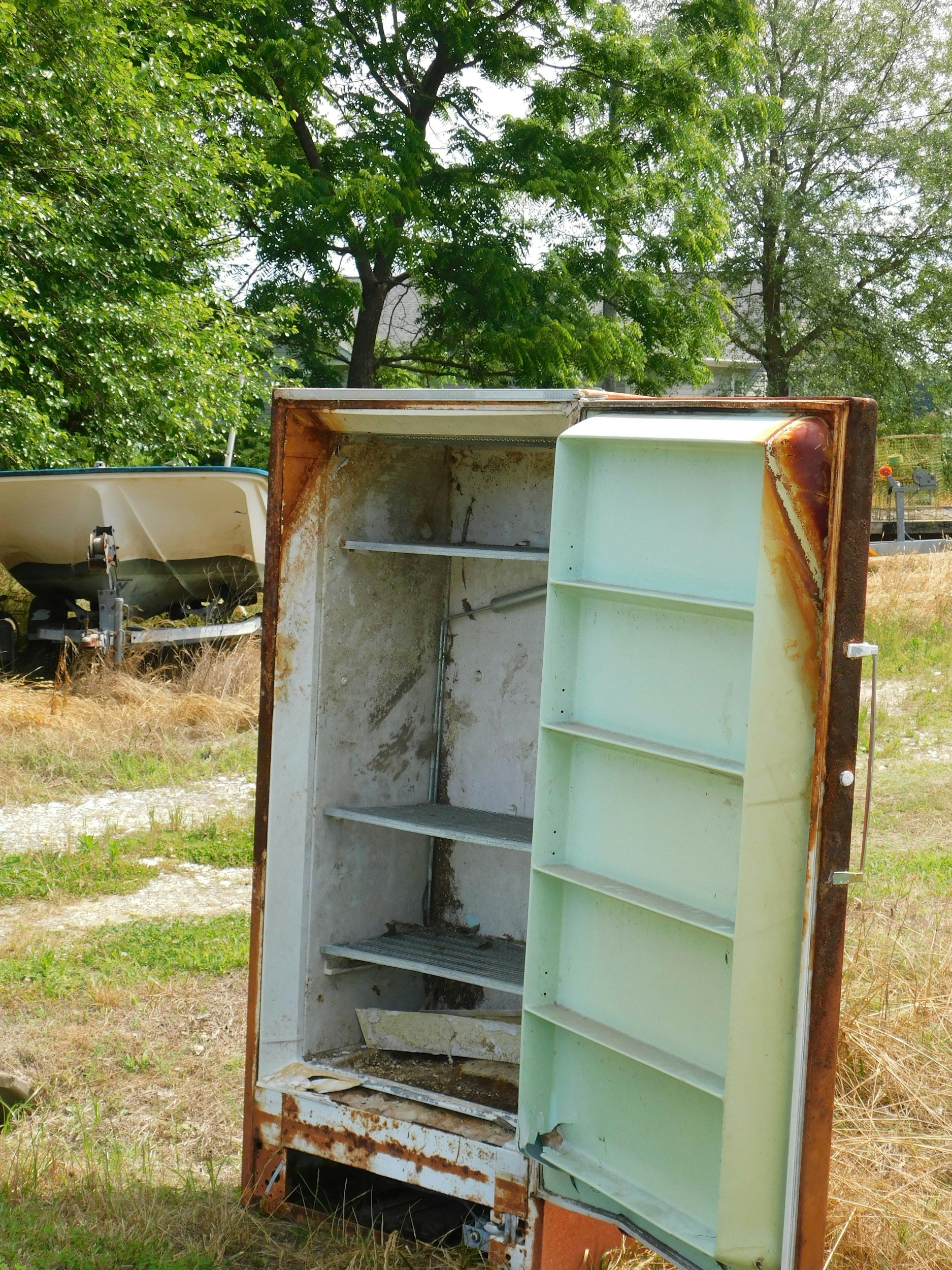 An old, rusty, and empty refrigerator outdoors with open doors, showing inside shelves and interior walls; background includes trees and another boat on a trailer.