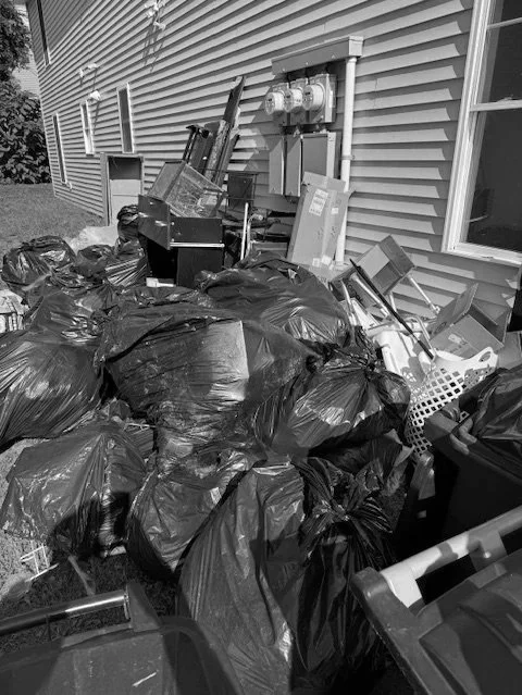 Pile of garbage bags and discarded household items outside a house with siding, near an electrical meter and outlet.