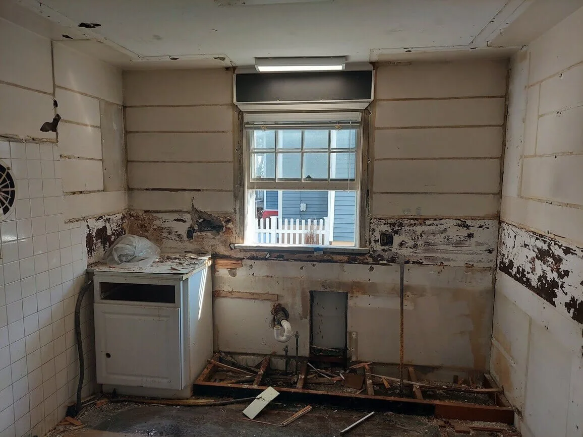 Room under renovation with damaged walls, exposed pipes, and debris on the floor.