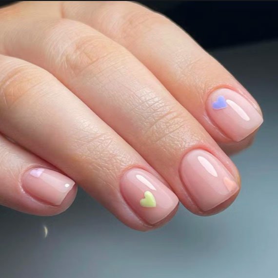 Close-up of a hand with manicured nails painted with clear polish; each nail features a small, pastel-colored heart or teardrop design, including purple, yellow, and green.