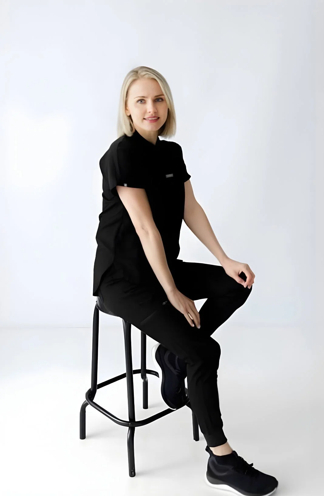 A woman with blonde hair wearing a black athletic outfit and black sneakers sitting on a black stool in front of a plain white background.