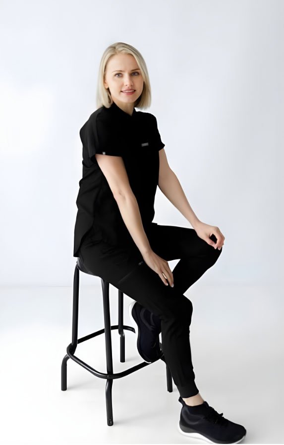 A woman with blonde hair wearing a black sports outfit sitting on a black stool against a white background.