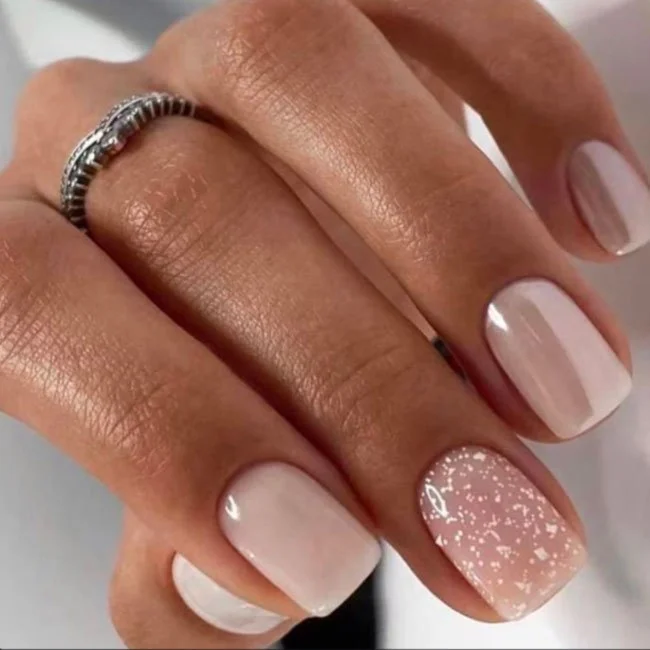 Close-up of a hand with manicured nails, featuring a light pink color and a glitter accent on the ring fingernail, and a silver ring on the middle finger.