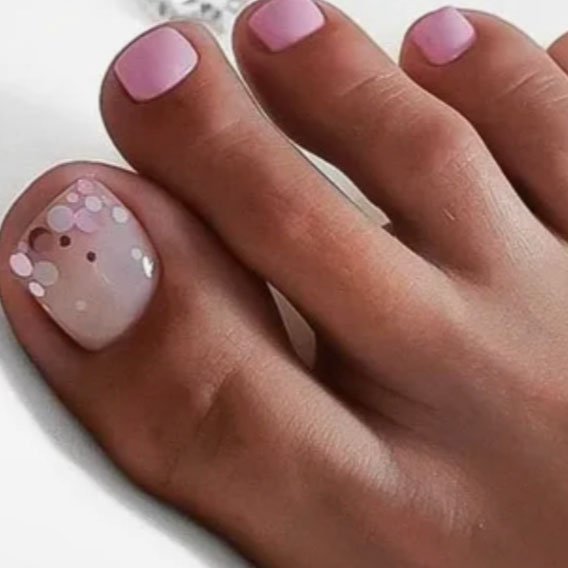 Close-up of toes with manicured nails, featuring pink nail polish on the second toe and a decorated big toe with pink and silver circular glitter accents.