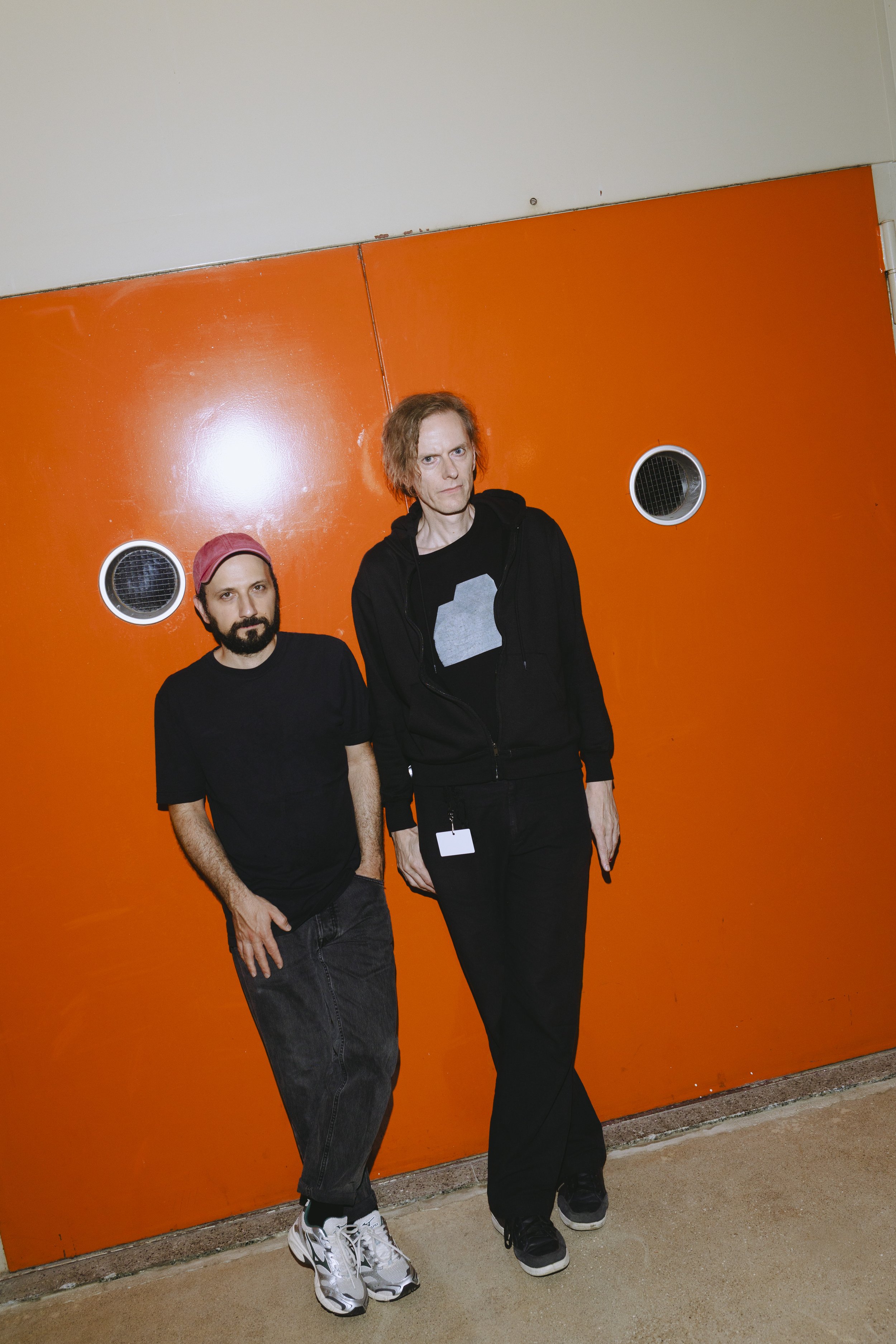 Zwei Männer stehen vor einer orangefarbenen Tür mit runden Fenstern.