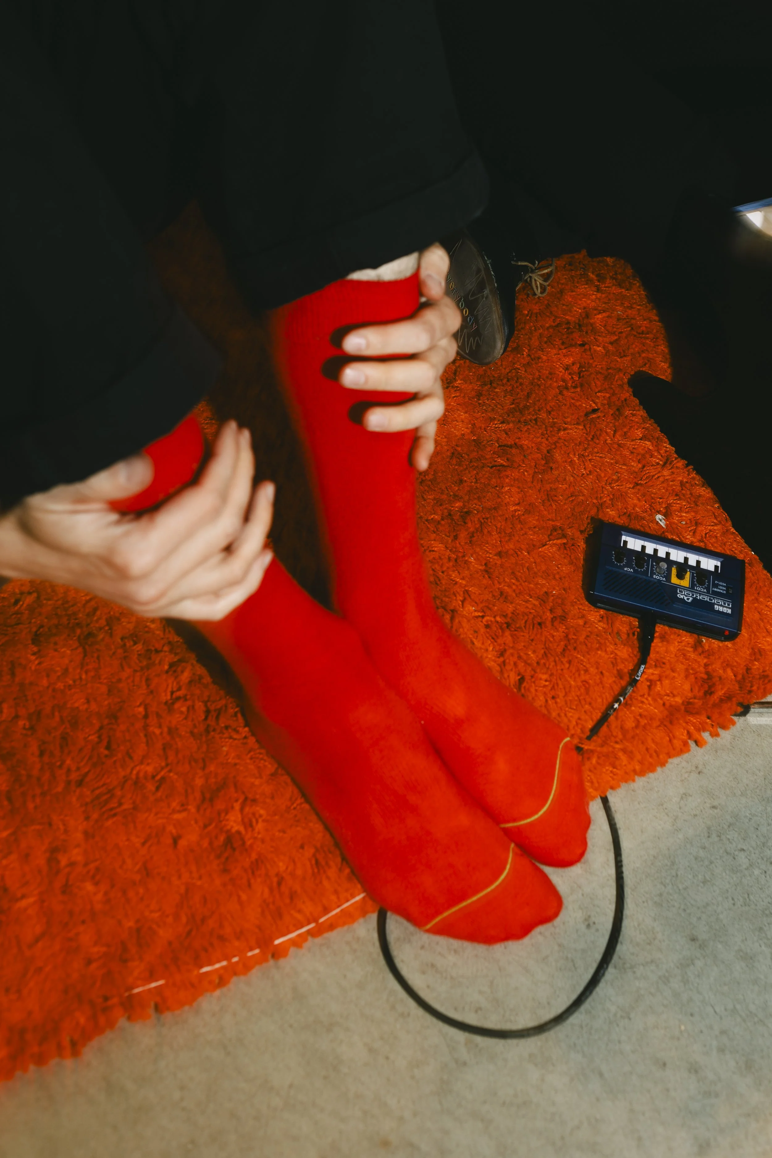 Personen, die rote Strümpfe trägt und  auf einem orangefarbenen Teppich sitzt, mit einem technischen Gerät daneben.