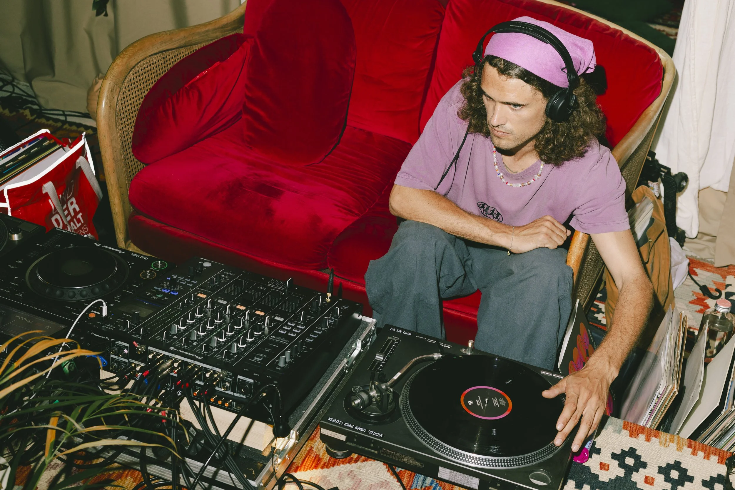 Ein Mann mit langen, lockigen Haaren und pinkem Kopfband, der Plattenspieler und DJ-Equipment bedient, sitzt auf einem roten Sofa in einem Raum mit buntem Teppich.