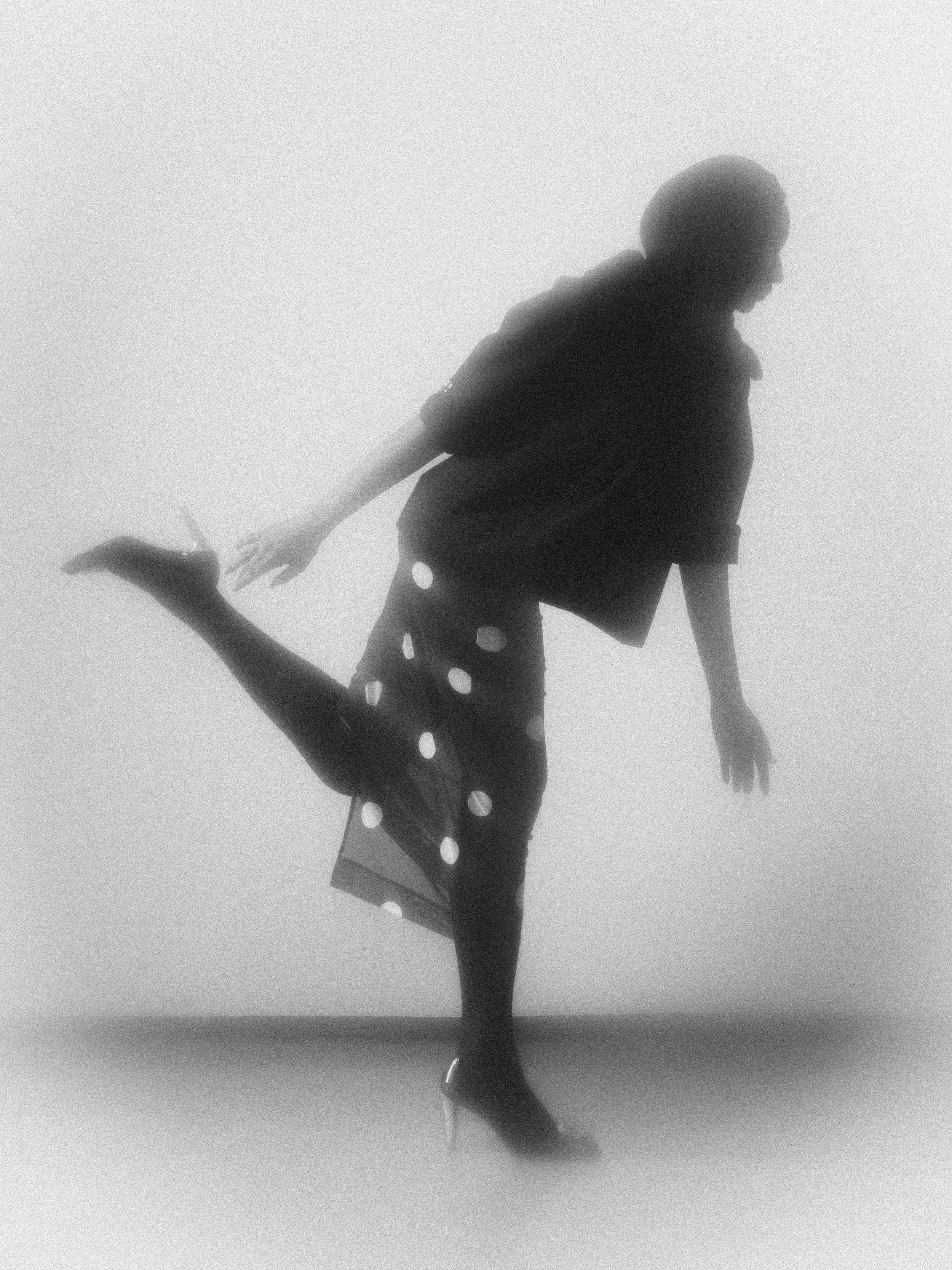 Schwarz-weiß-Fotografie einer Person, die auf einem Bein balanciert, mit einem Arm nach hinten gestreckt, in einem Nebel- oder Rauchstil, die Kleidung mit Punkten trägt.