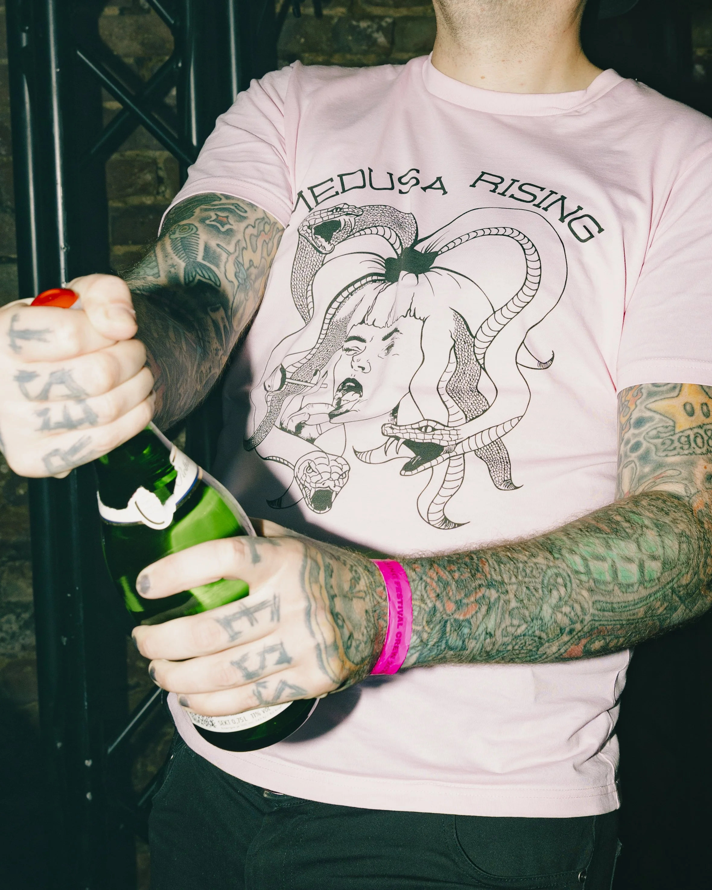 Nahaufnahme einer Person in einem rosa T-Shirt mit grafischem Druck und Tätowierungen auf Armen, die eine grüne Sektflasche hält.